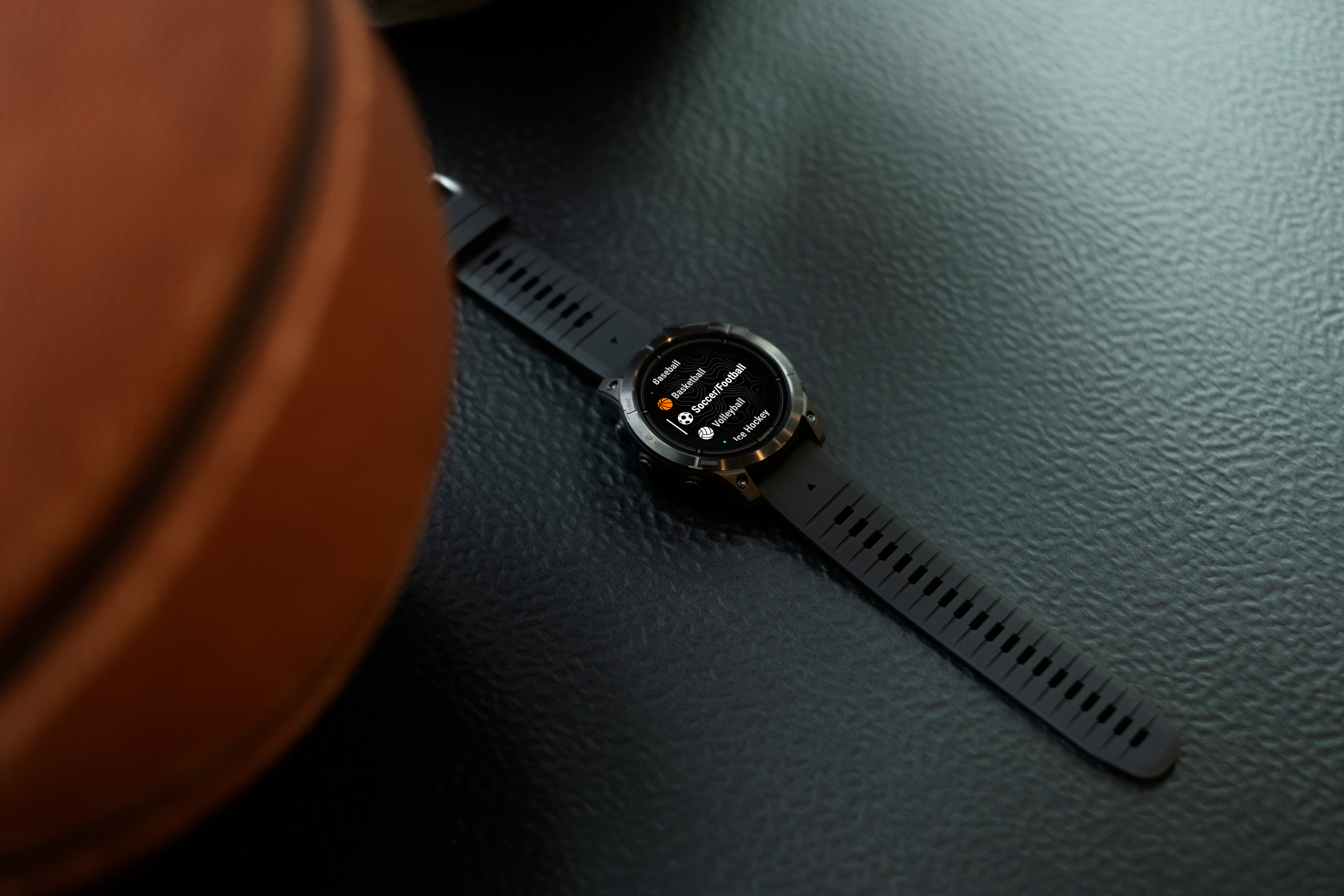 Une montre intelligente noire affiche des options sportives comme le baseball et le football. Un ballon de basket est partiellement visible en arrière-plan.