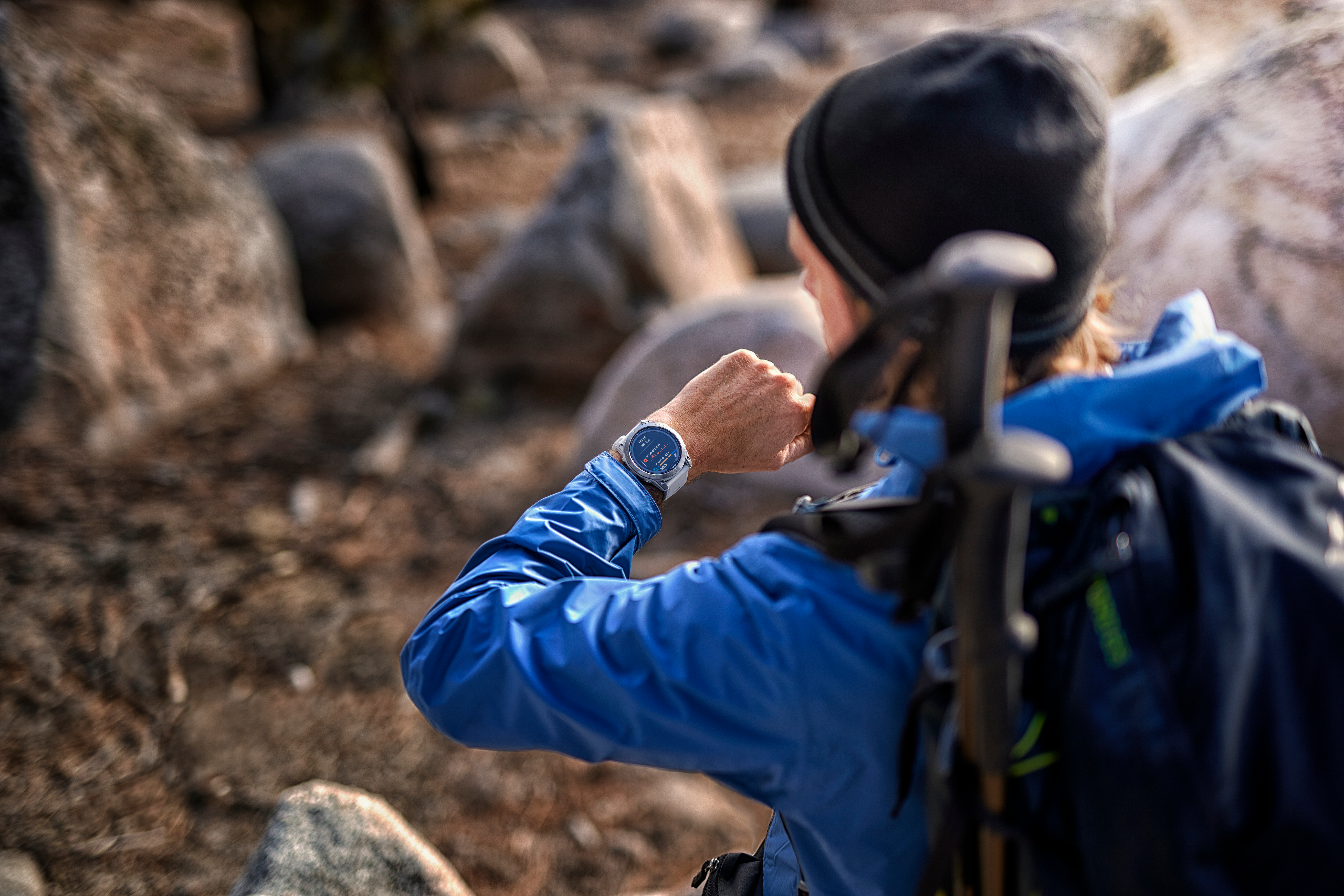 Eine Person in blauer Jacke und schwarzem Hut schaut auf eine Uhr. Felsen im Hintergrund. Wanderstöcke sind sichtbar.