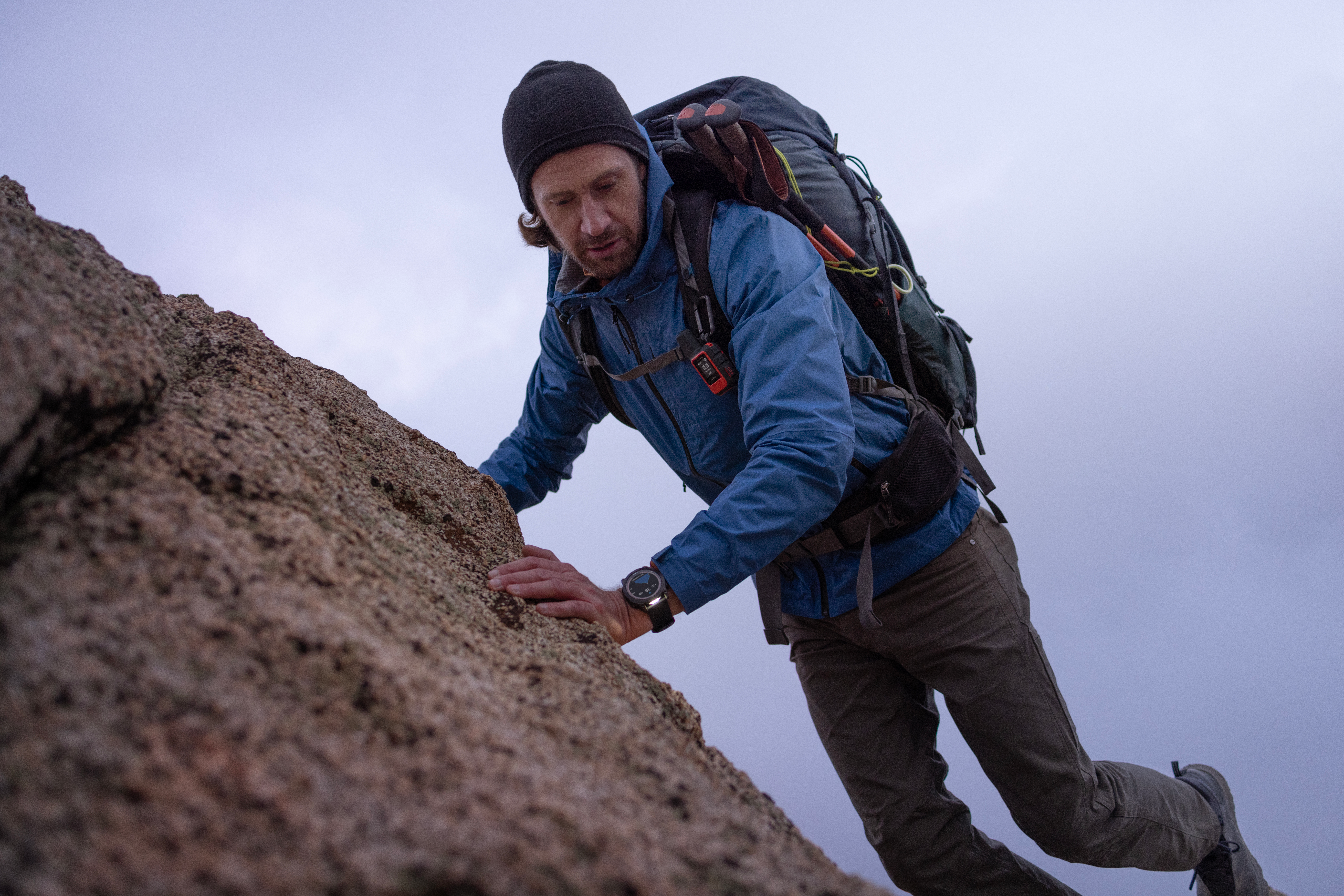 Ein Mann klettert eine Felswand hinauf. Er trägt eine blaue Jacke, eine schwarze Mütze und einen Rucksack. Der Himmel ist bedeckt.