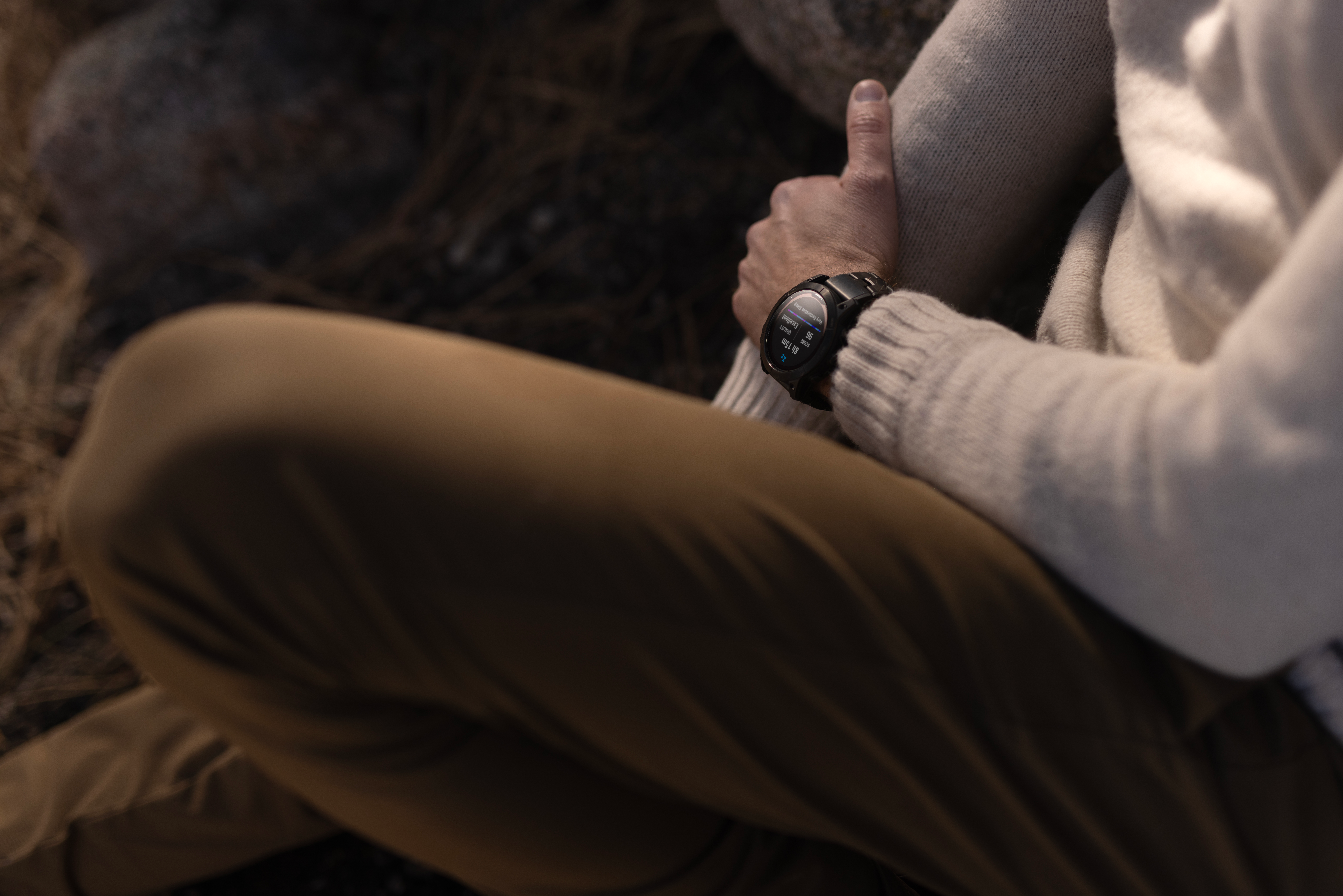 Une personne portant une montre intelligente est assise. Le cadran affiche des données de sommeil. La personne porte un pull blanc et un pantalon marron.