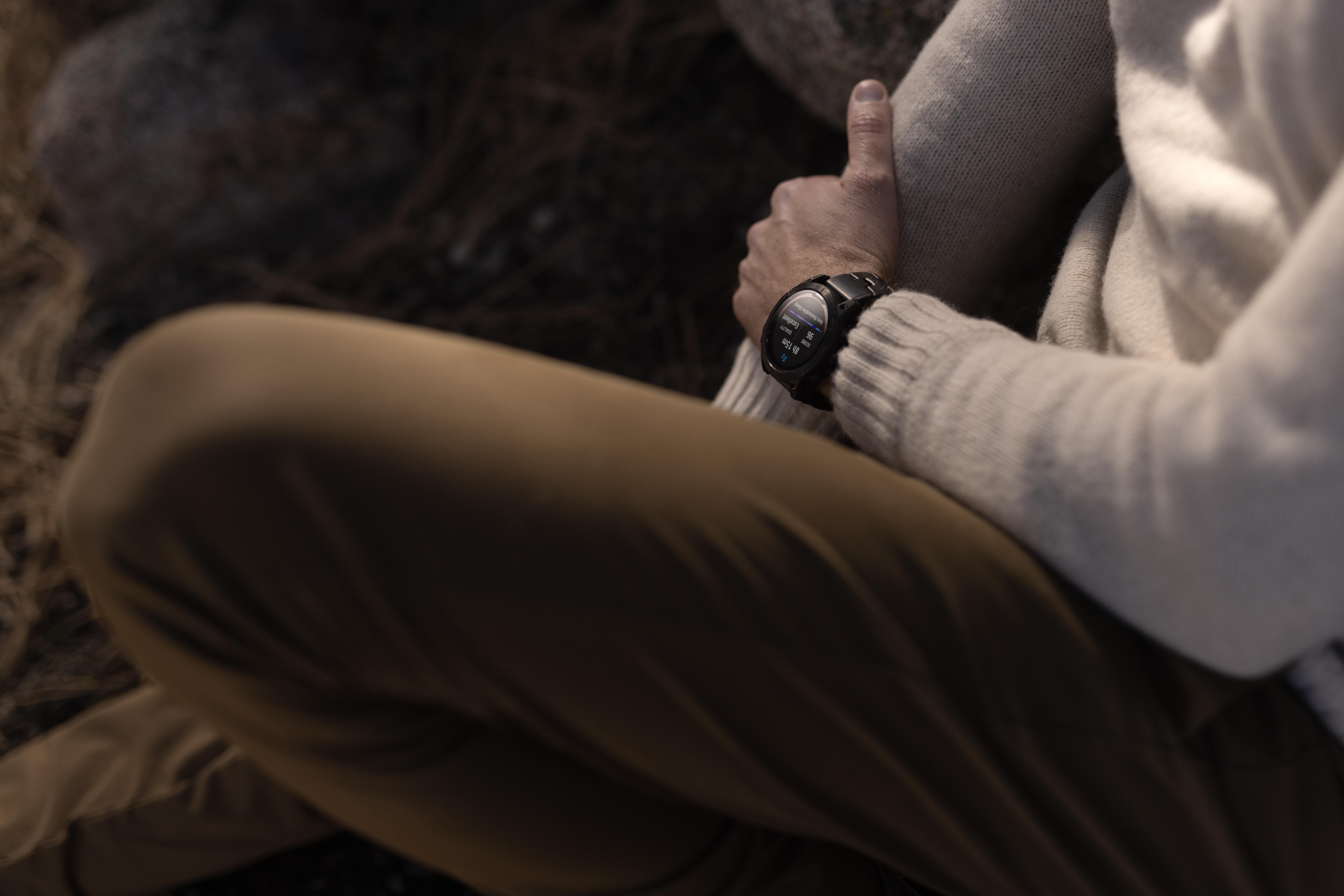 Person mit schwarzer Smartwatch, sitzend mit verschränkten Beinen, Hände im Schoß gefaltet.