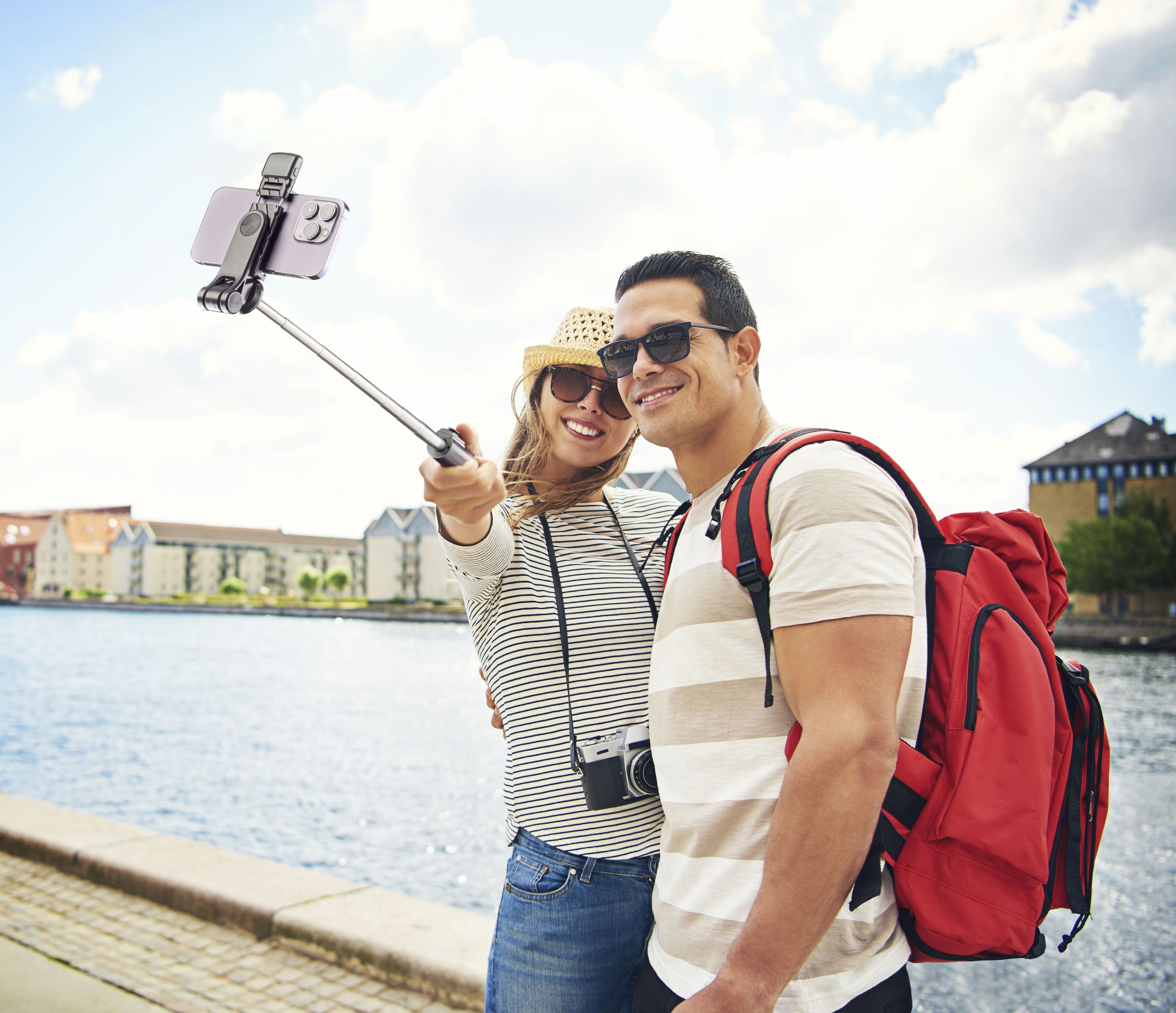 Pareja tomando selfie con teléfono en un palo selfie. Agua y edificios fondo.
