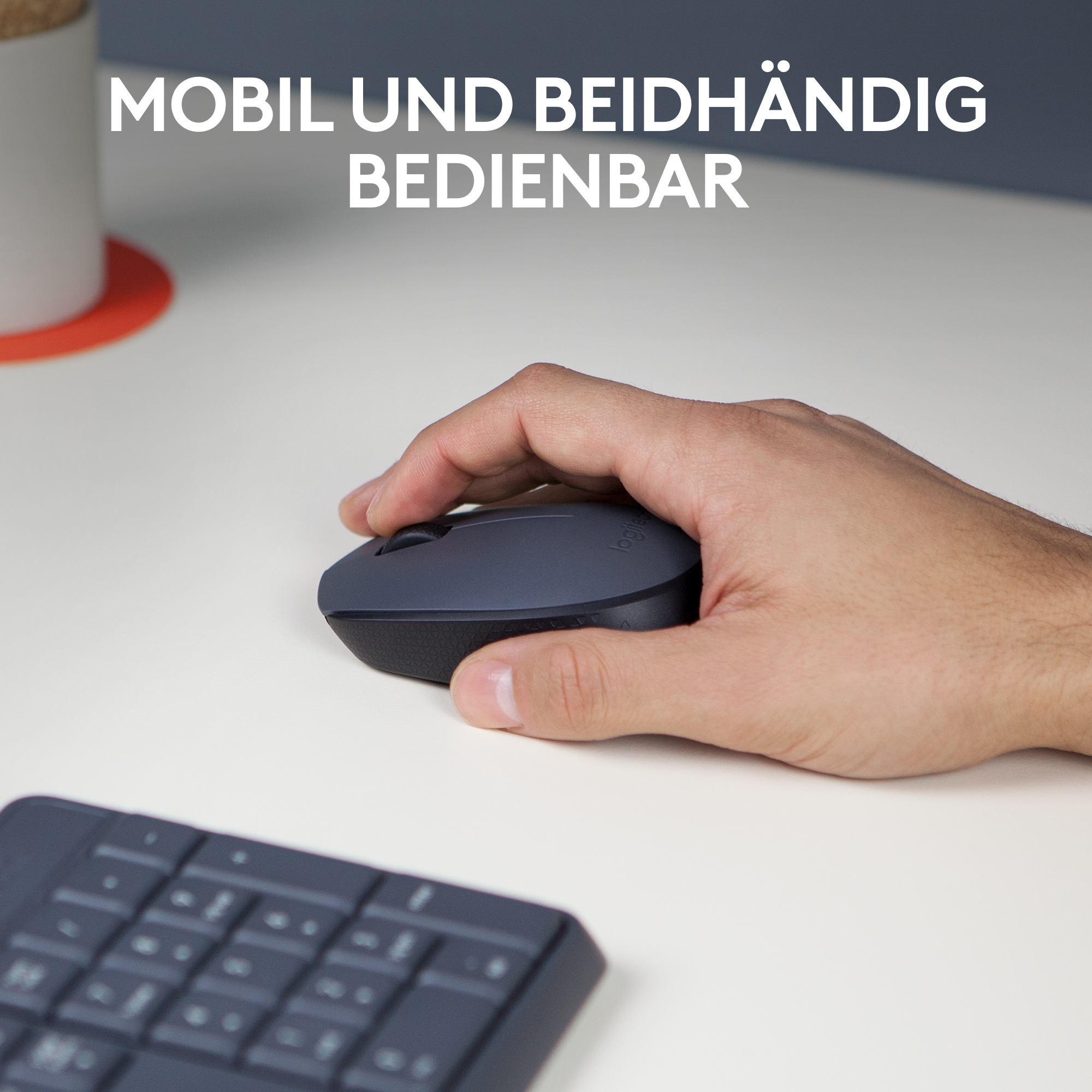 Eine Hand hält eine graue Maus auf einem weißen Schreibtisch, im Hintergrund eine Tastatur.