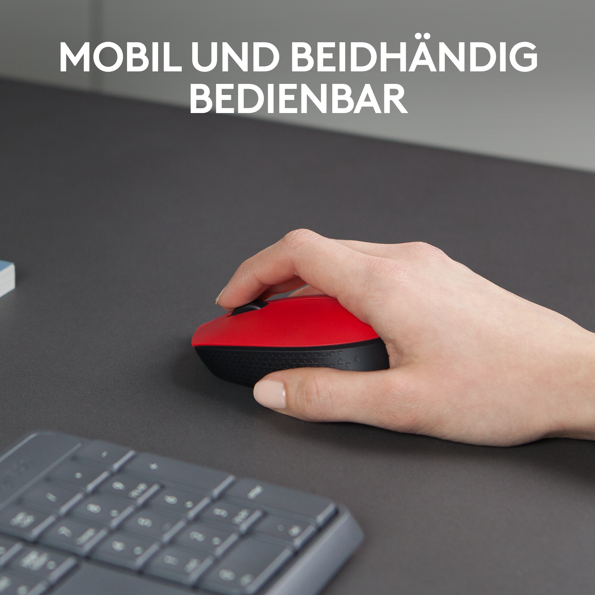 Die Hand einer Person, die eine rote Maus auf einem grauen Schreibtisch hält, mit Tastatur.