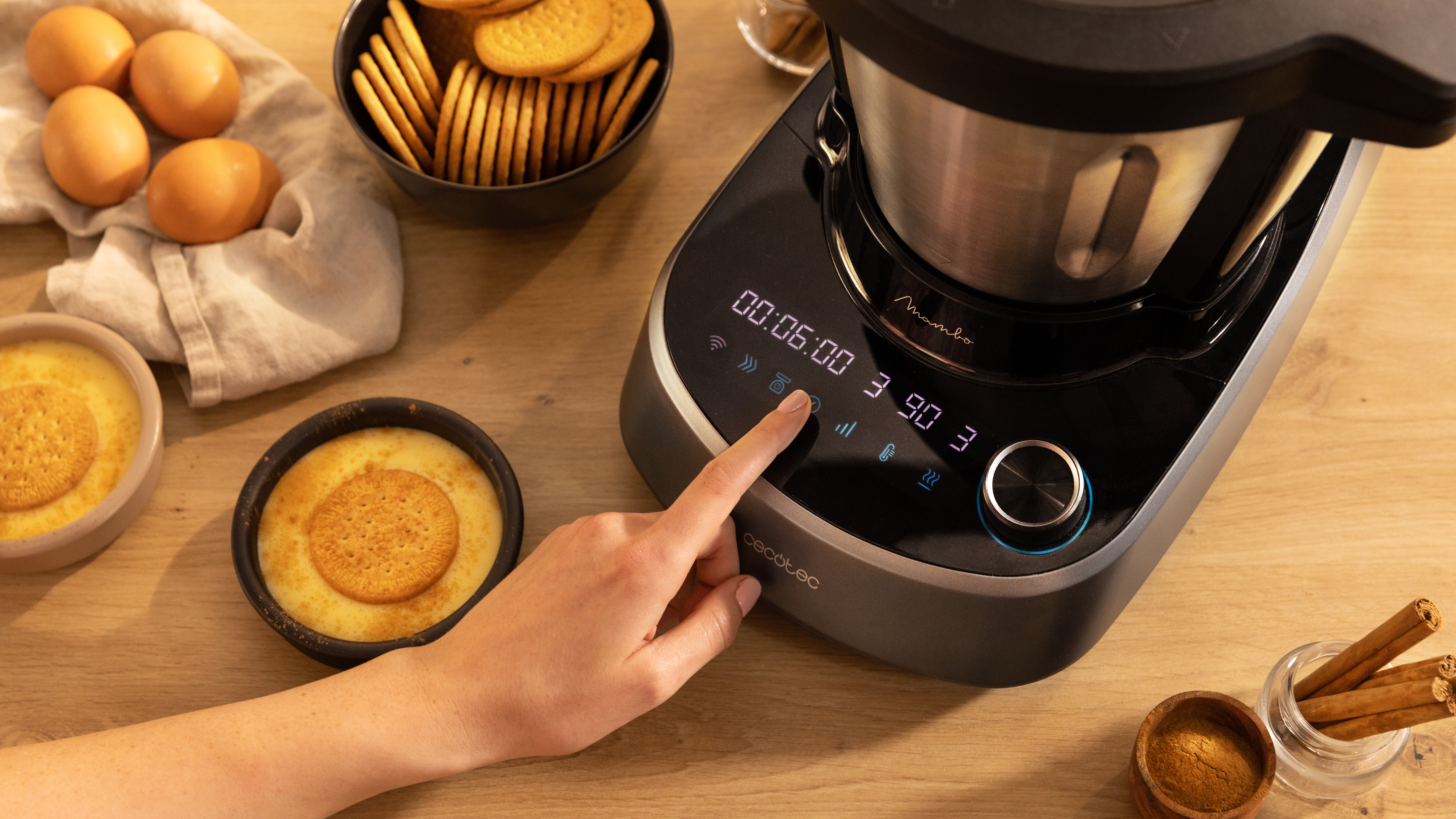 Una mano toca una máquina de cocina con una pantalla digital. Incluye un cuenco de galletas y huevos.