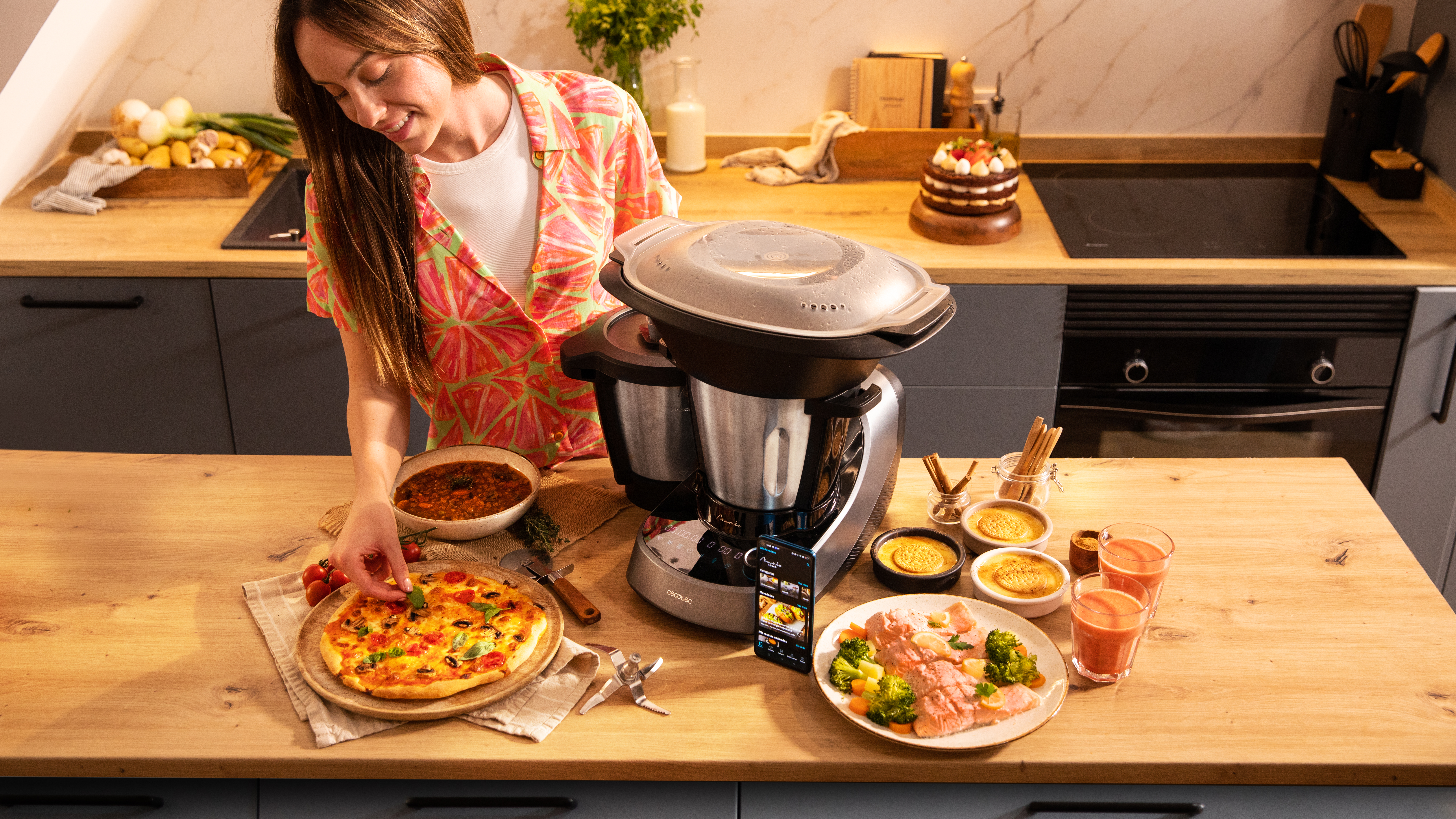 Una mujer en una cocina usa un procesador de alimentos. Una pizza y ensalada están en la encimera.