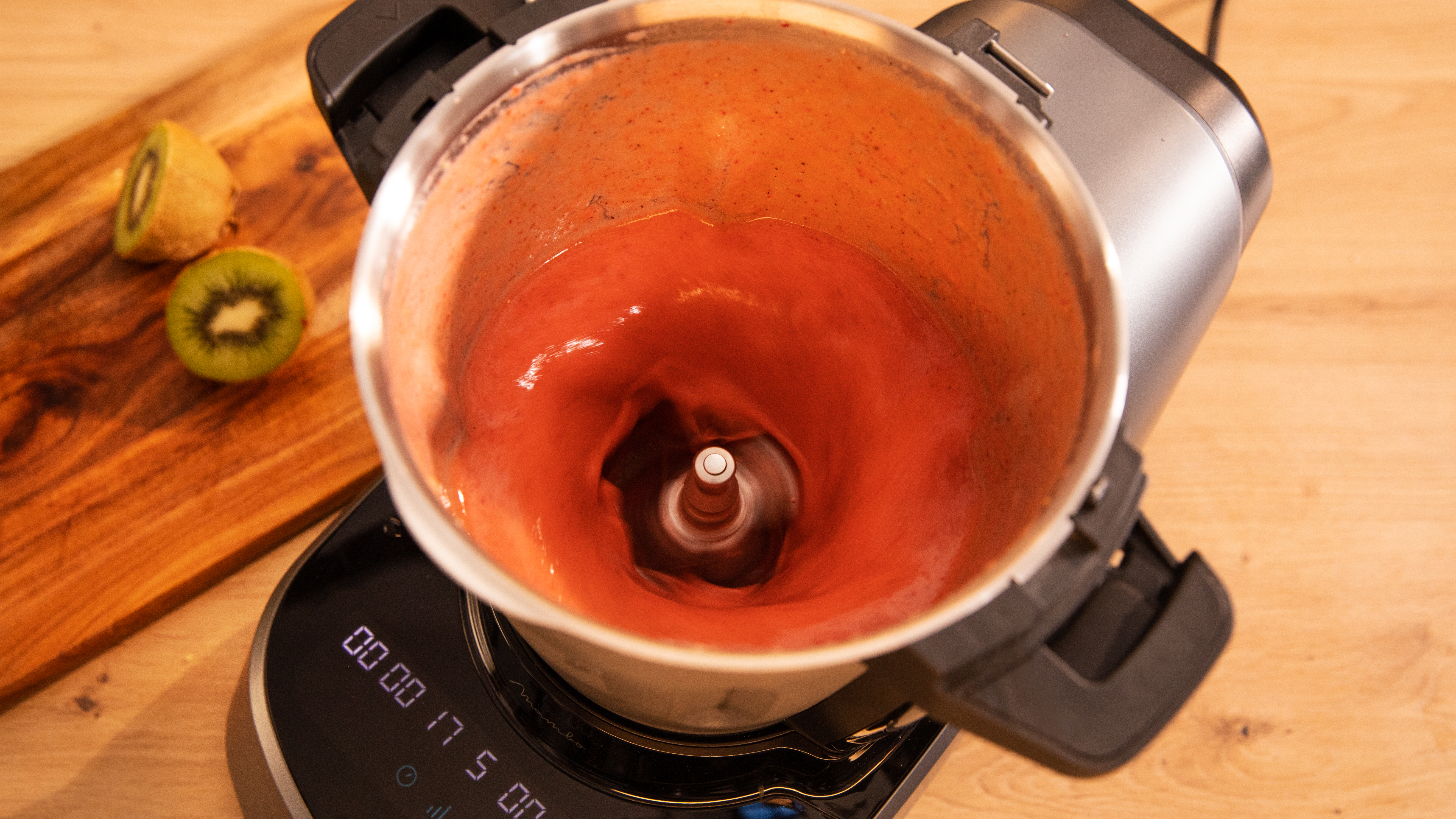 Un procesador de alimentos mezclando un batido rojo. Rodajas de kiwi a un lado.