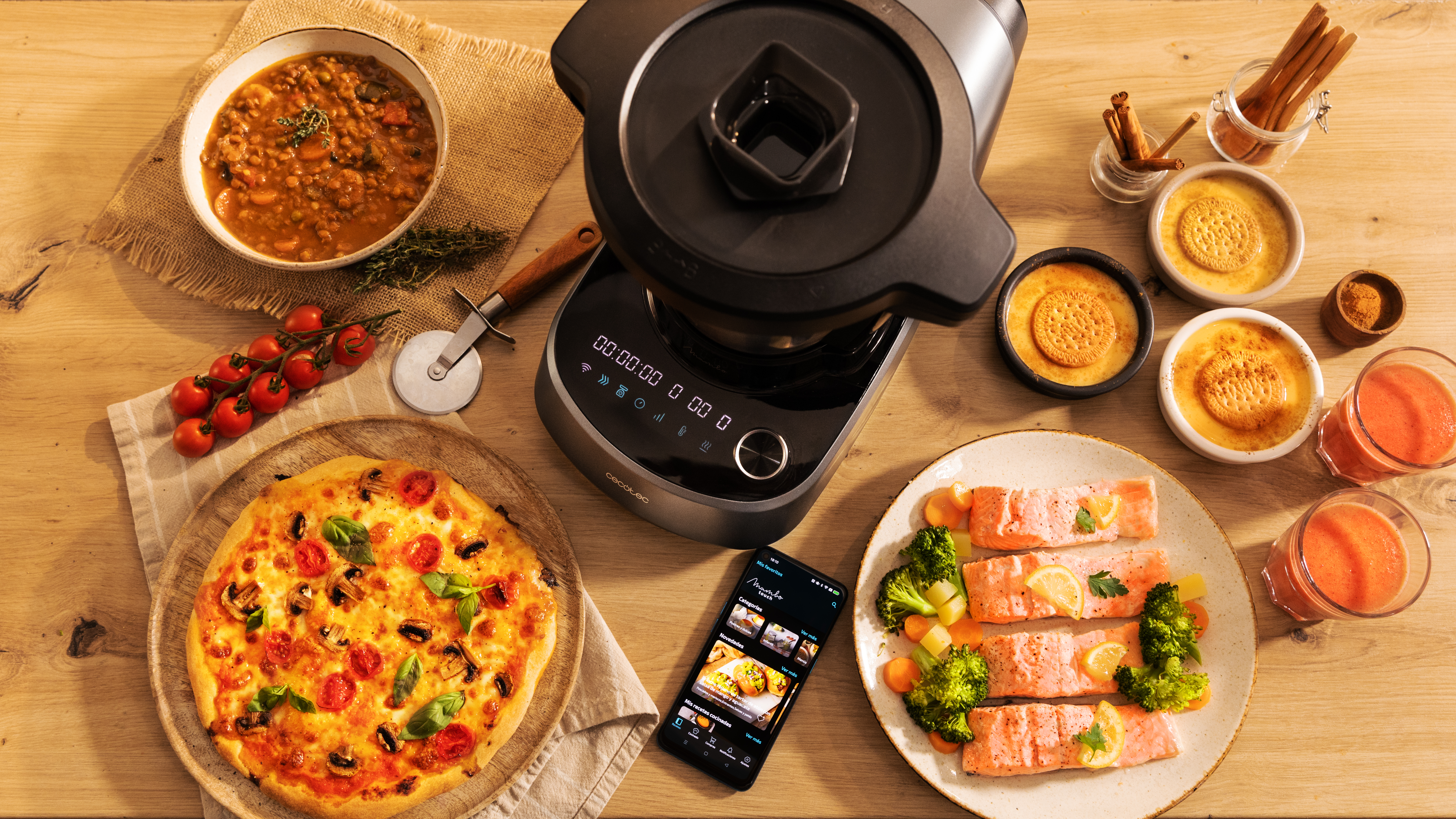 Una mesa con una multicocina, comida y un smartphone. Hay pizza, salmón y postres.