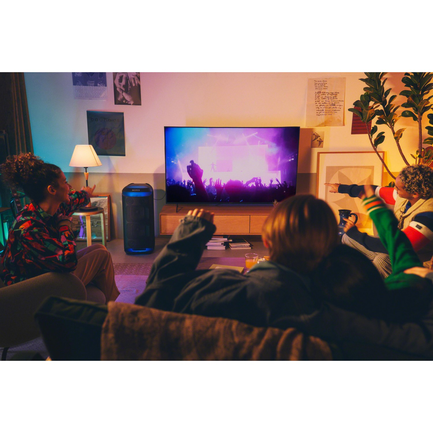 Un groupe de personnes regardant la télé, un grand haut-parleur et une lampe sont également présents.