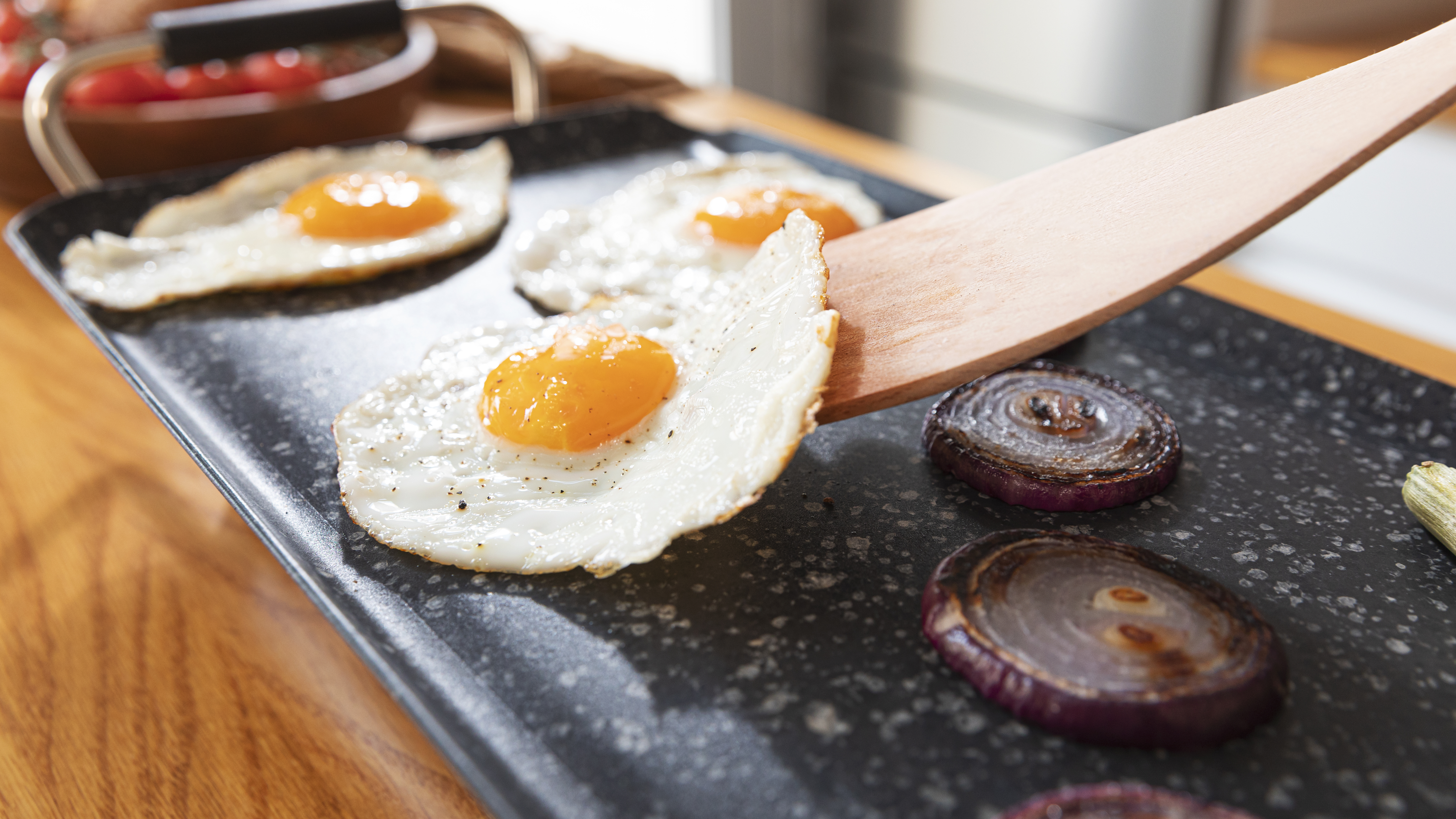 Huevos cocinándose en una plancha negra con cebollas rojas. Espátula de madera en la escena.