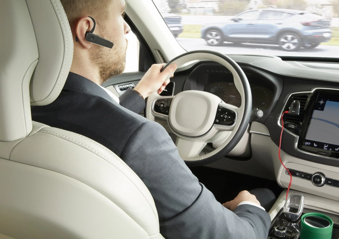 Un homme en costume conduit une voiture, portant un casque Bluetooth. Une autre voiture en arrière-plan.