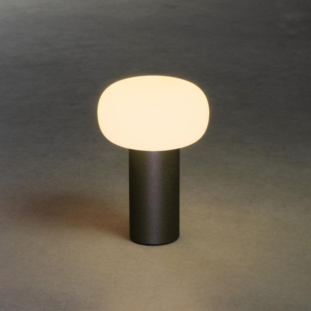 Eine moderne Lampe mit dunkelgrauem Sockel und einem ovalen, leuchtenden weißen Schirm, auf grauer Oberfläche.