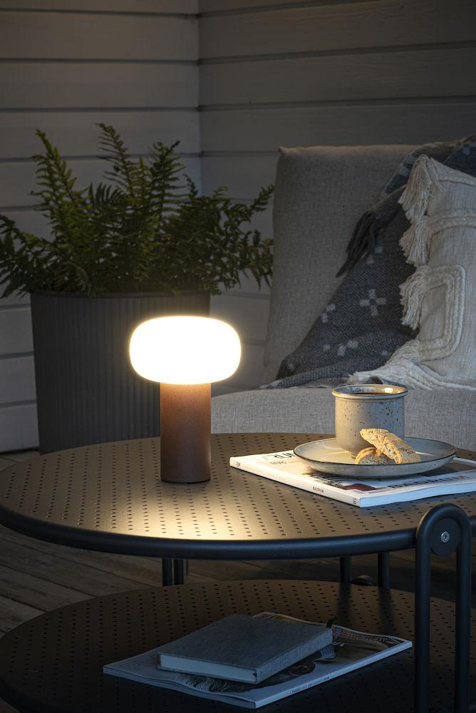 Eine beleuchtete Lampe auf einem Tisch neben einer Pflanze, einer Zeitschrift und einer Tasse. Die Szene ist eine Terrasse.