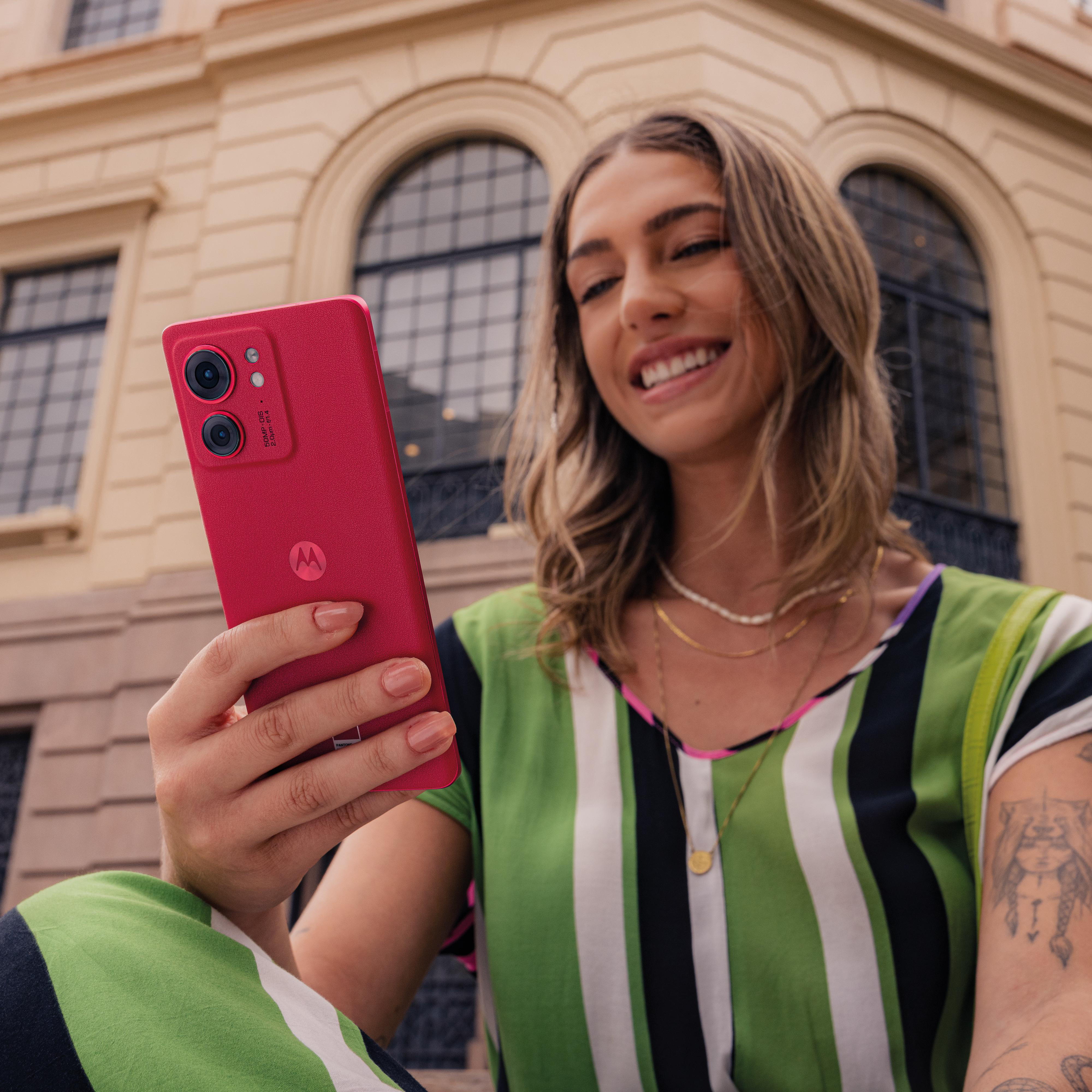 Frau hält ein rotes Smartphone, lächelt. Im Hintergrund ein Gebäude mit Bogenfenstern.