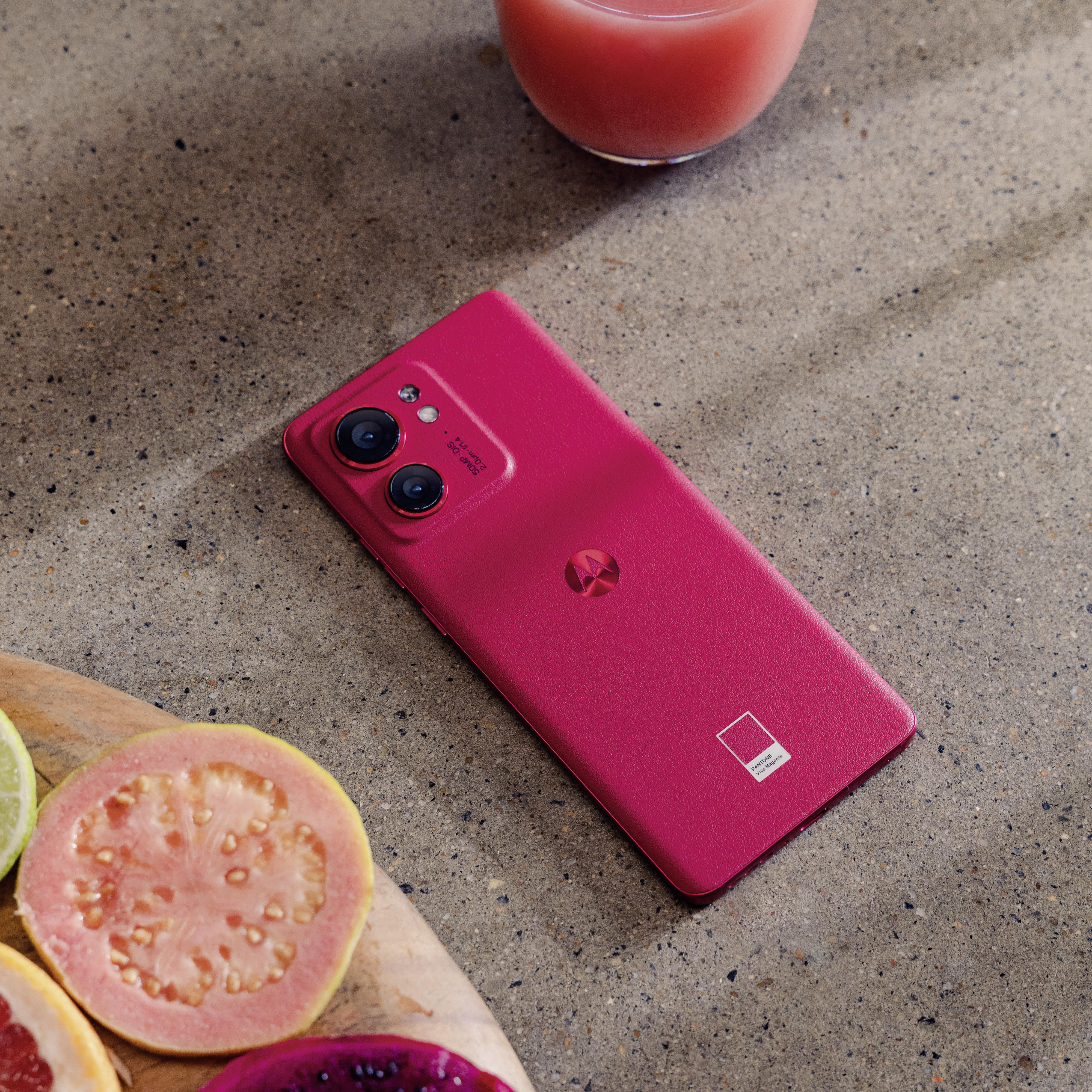 Rosafarbenes Motorola-Telefon auf einem Tisch mit Obst und einem Getränk. Das Telefon hat zwei Kameras.