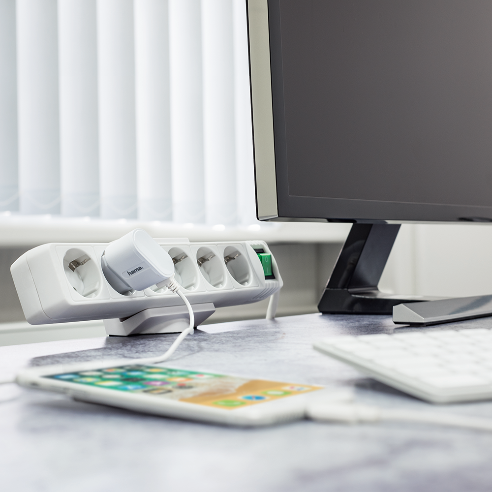 Witte stekkerdoos met een oplader aangesloten. Telefoon en monitor zichtbaar op de achtergrond op een bureau.