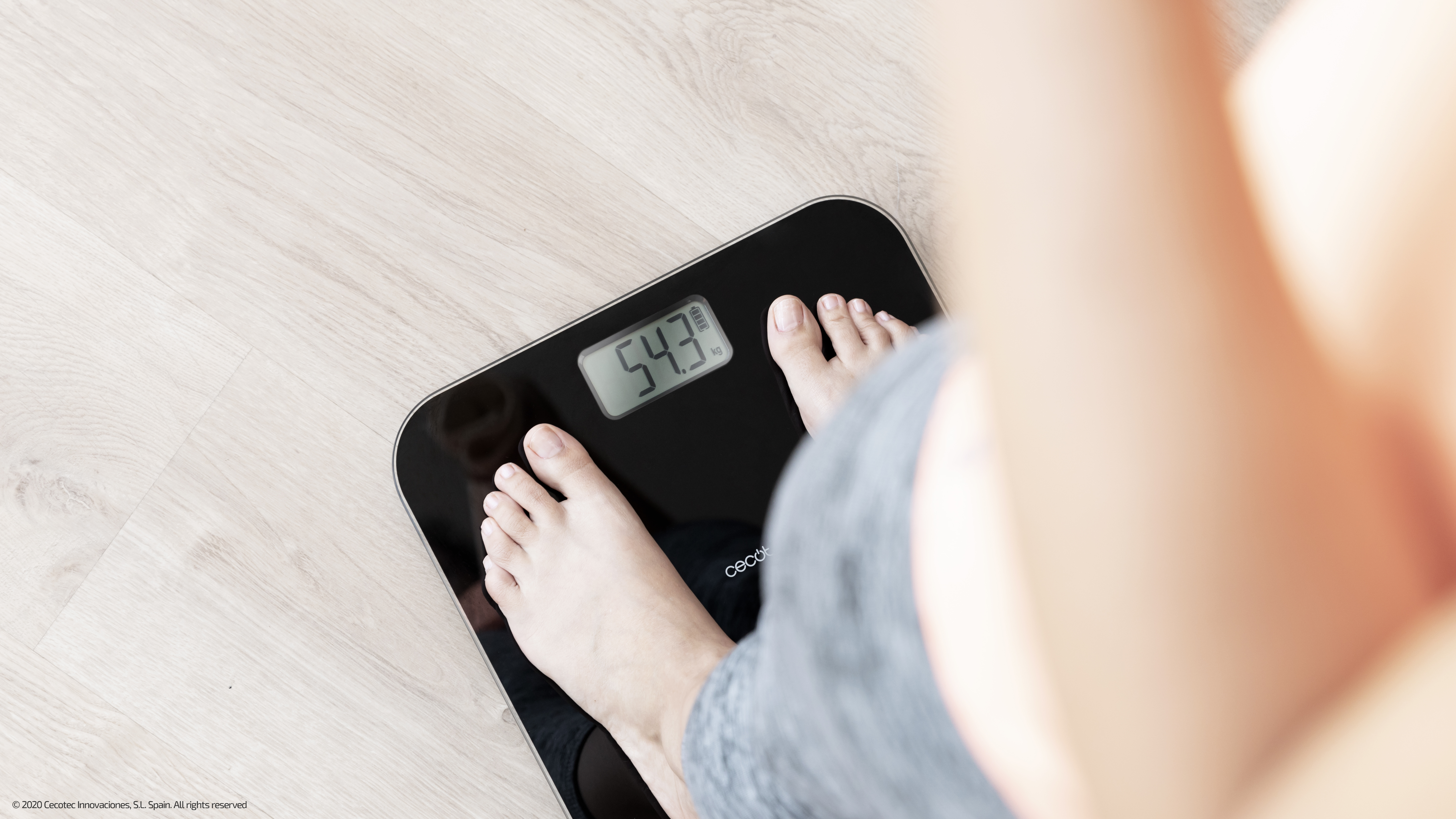 Pies en una báscula digital negra. Muestra 54.3 kg. Suelo de madera clara.