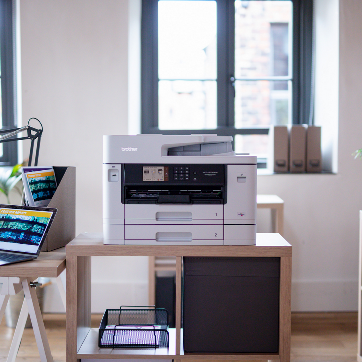 A white and gray printer on a wooden desk with a laptop, desk organizer and file folders. Window in background.