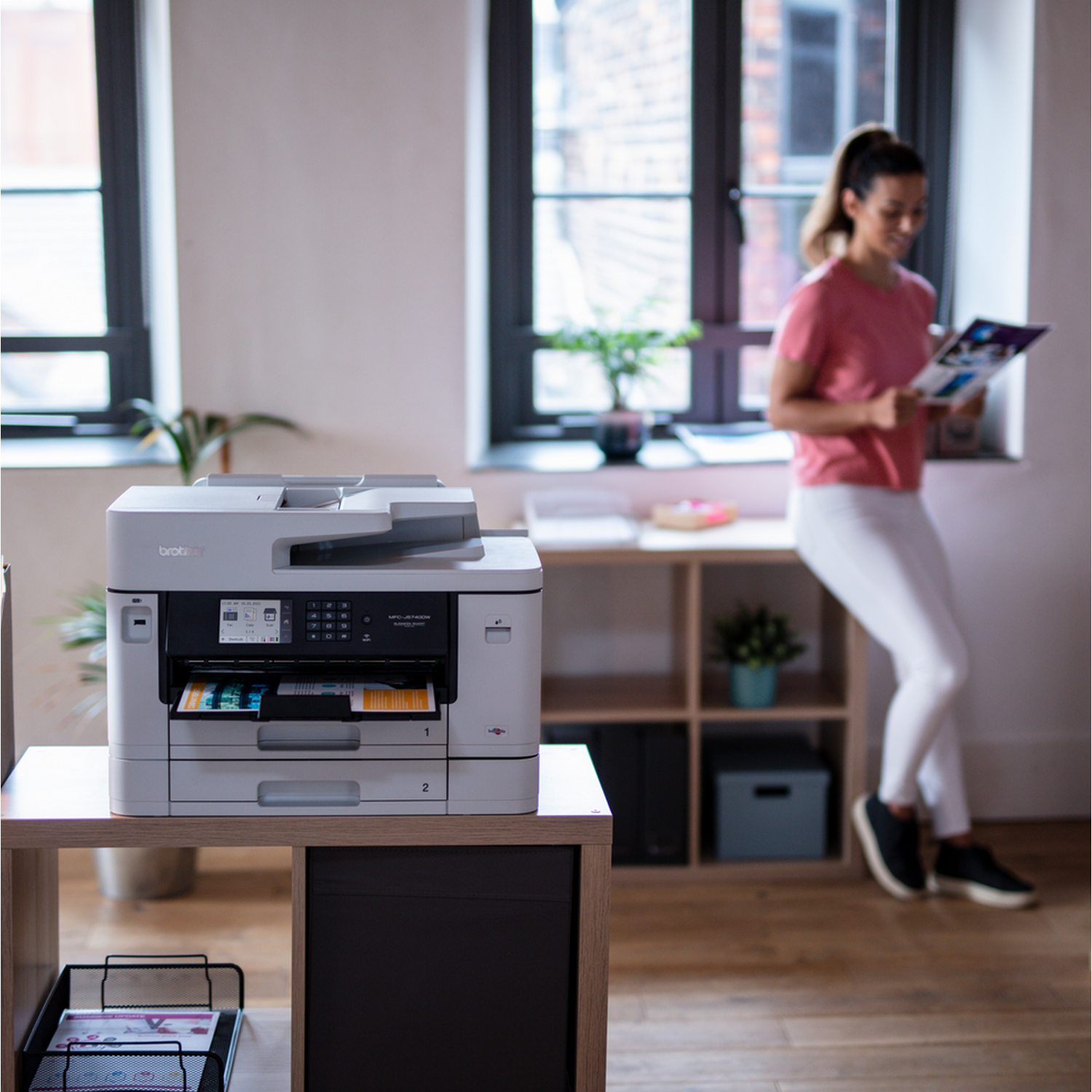 A printer on a table with a woman in the background. The printer is white and grey.