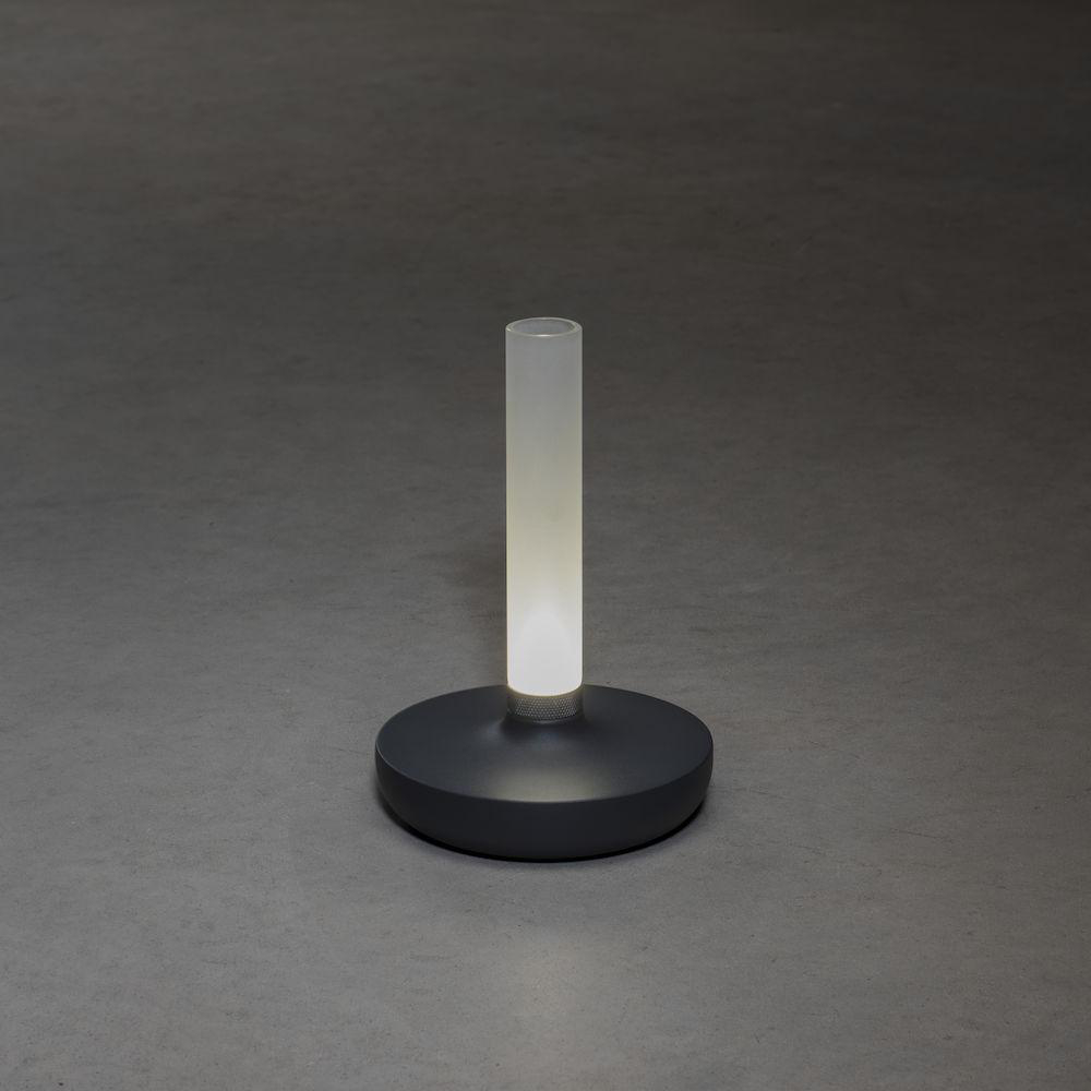 Eine hohe, zylindrische Lampe mit weißem Licht. Dunkelgrauer Sockel auf grauer Oberfläche.