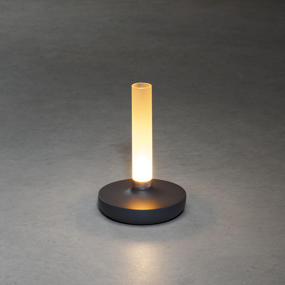 Graue Lampe mit zylindrischer Lichtquelle auf dunkelgrauer runder Basis, steht auf grauer Oberfläche.