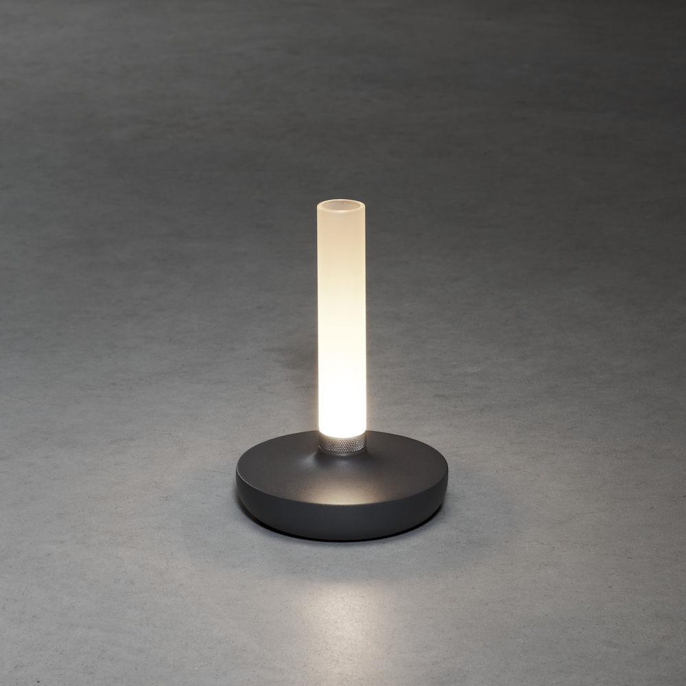 Eine beleuchtete zylindrische Lampe auf einem dunkelgrauen runden Sockel auf einer grauen Oberfläche.