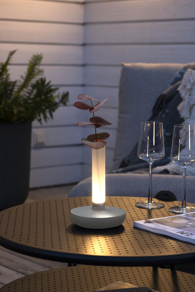 Ein Tisch mit einer beleuchteten Lampe mit Blättern, zwei Weingläsern und einer Zeitschrift, vor verschwommenem Hintergrund.