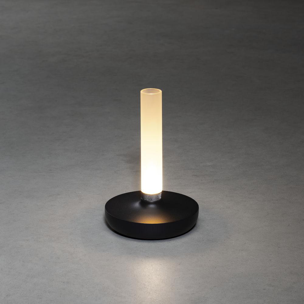Eine schwarze Tischlampe mit einem vertikalen, beleuchteten Rohr auf einem runden Sockel. Auf grauer Oberfläche.