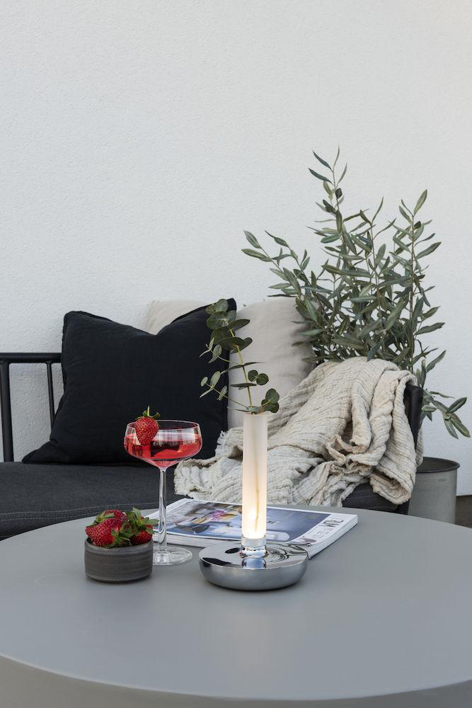 Ein Tisch mit Getränk, Erdbeeren und einer brennenden Kerze. Decke und Pflanze im Hintergrund.