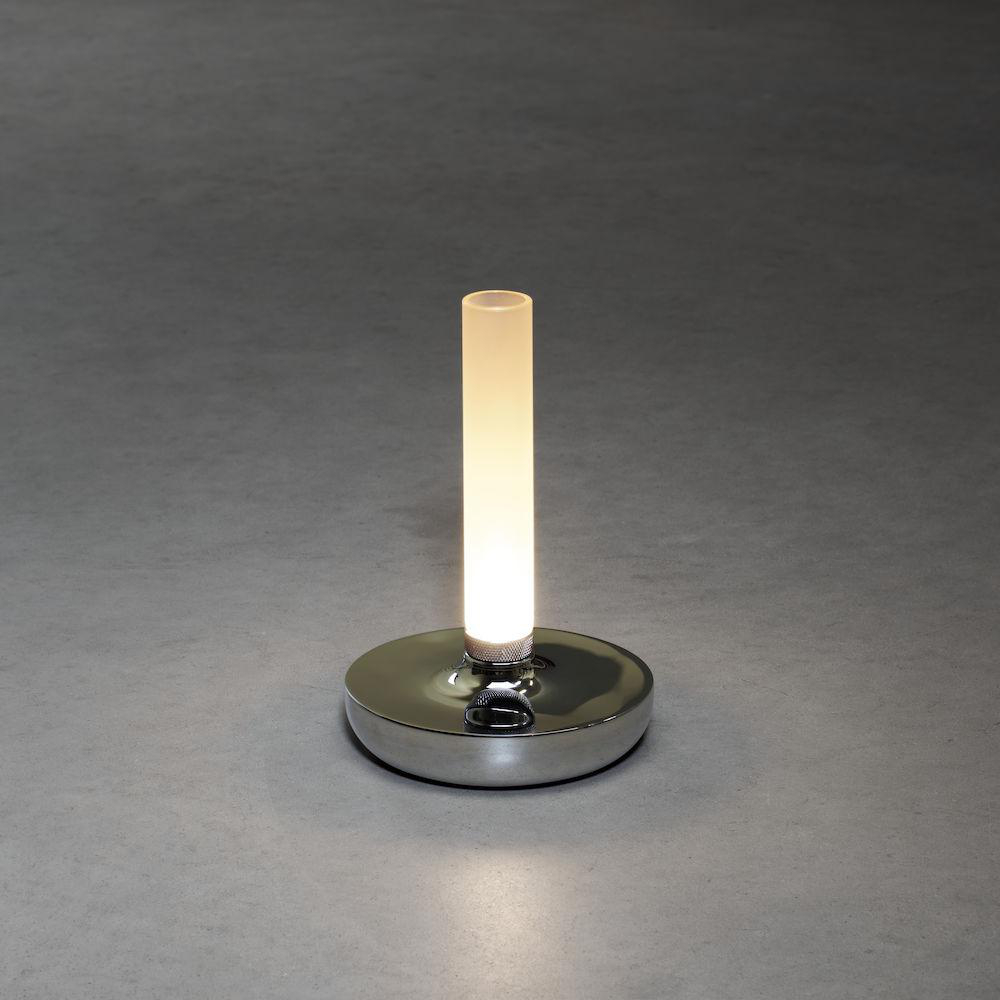 Eine silberne Lampe mit einem leuchtenden vertikalen Rohr auf einem runden Sockel, auf grauer Oberfläche.