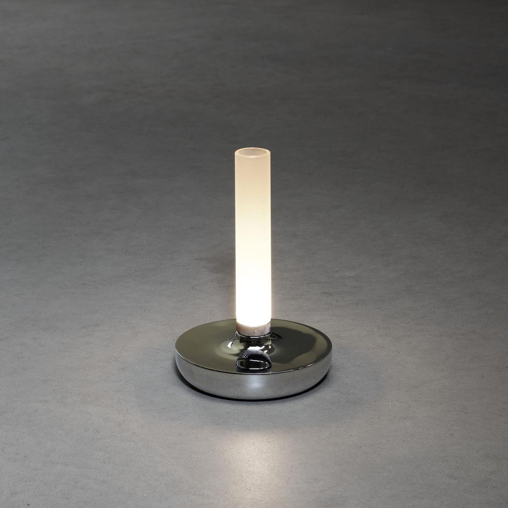 Eine silberne Lampe mit zylindrischem Licht auf rundem Sockel. Der Sockel ist reflektierend, auf grauer Oberfläche.