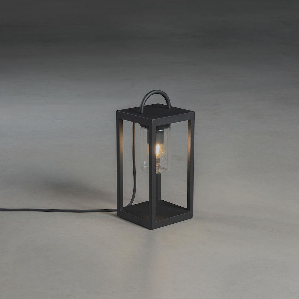 Eine schwarze Laterne mit einer Glühbirne, auf einer grauen Oberfläche. Ein schwarzes Kabel verläuft.