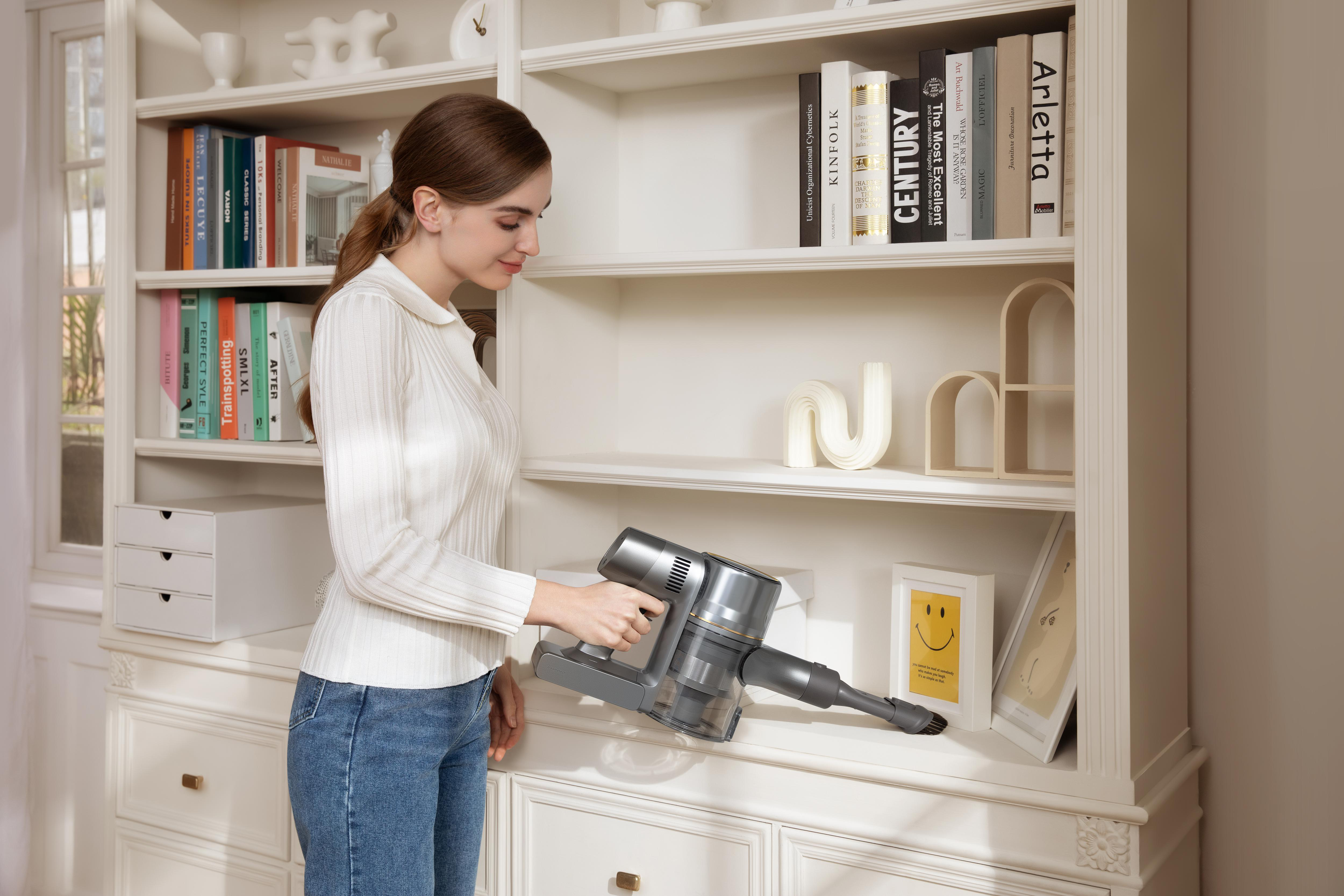 Eine Frau, die einen Handstaubsauger benutzt, um ein Regal in einem weißen Schrank mit Büchern und Dekorationen zu reinigen.