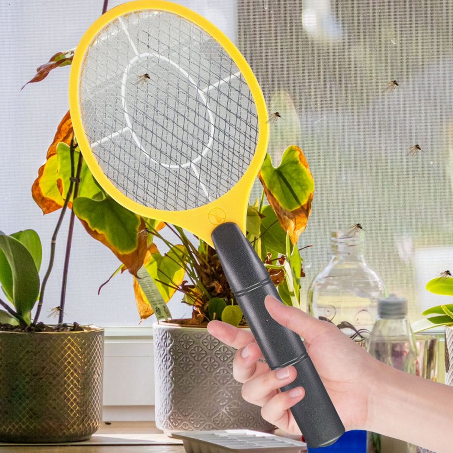 Hand die een gele elektrische vliegenmepper vasthoudt, met raam en vliegende insecten op de achtergrond.