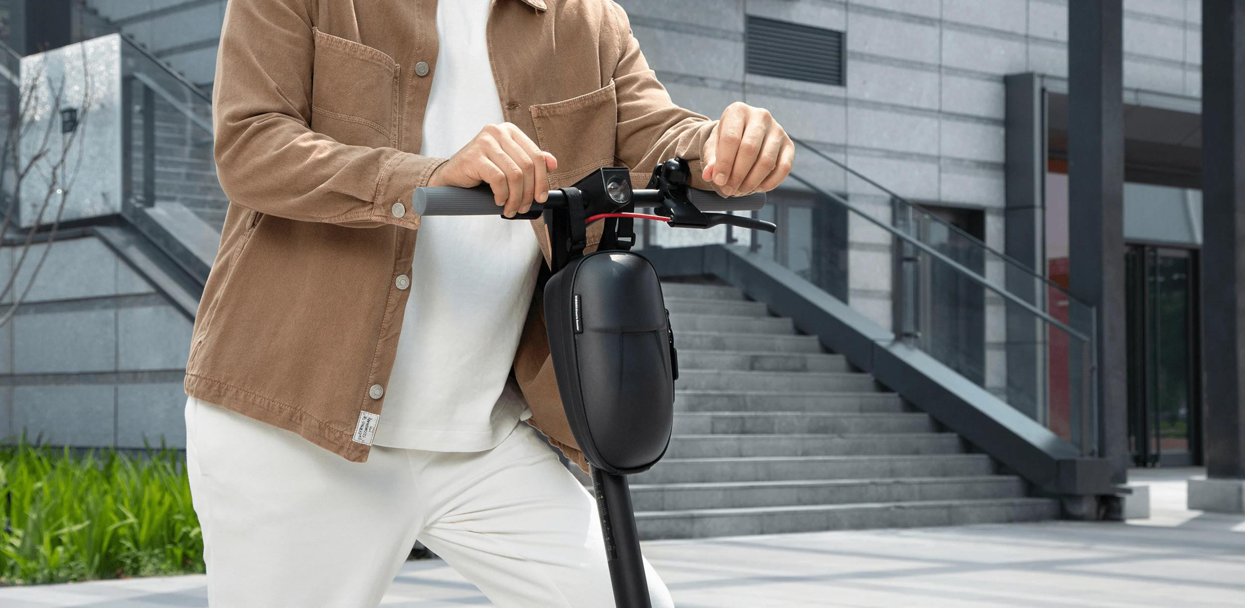 Mann hält einen Elektroroller. Trägt eine braune Jacke und weiße Hose. Urbaner Hintergrund.