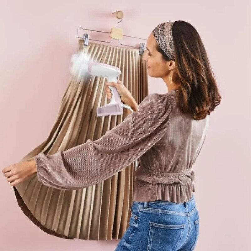 Kobieta parująca plisowaną spódnicę za pomocą parownicy ręcznej, różowe tło.