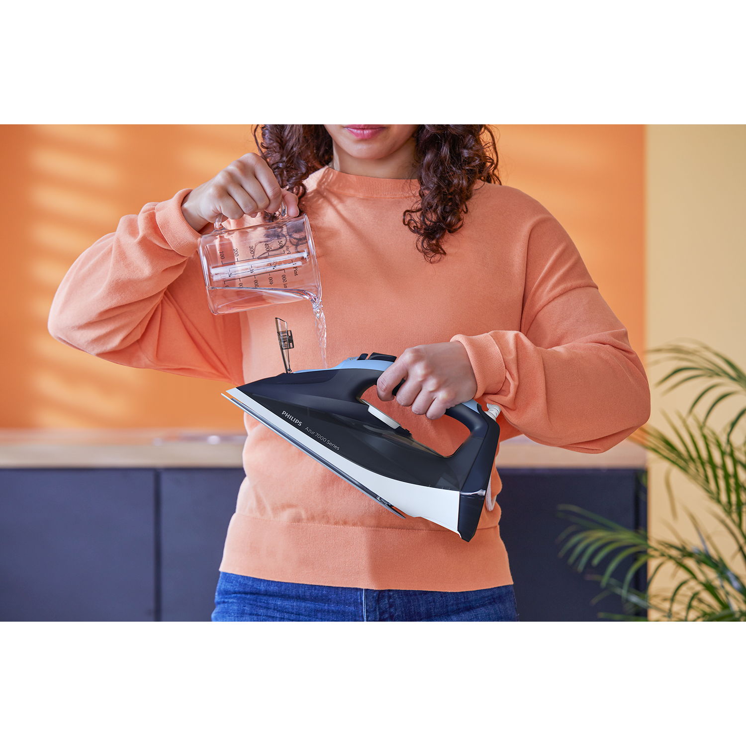 Woman pouring water into an iron, with orange background and black cupboards.
