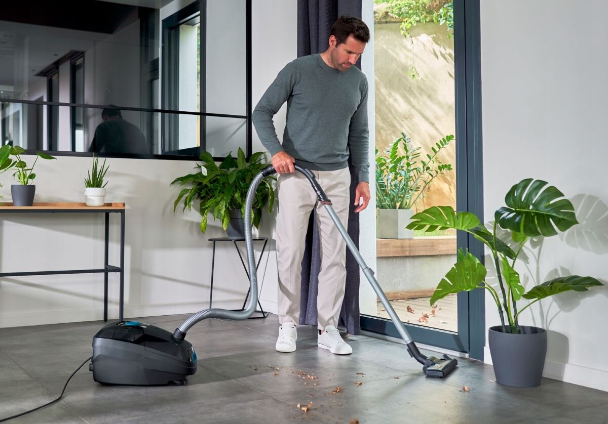 Mężczyzna odkurza pokój. Rośliny w tle. Podłoga jest brudna.