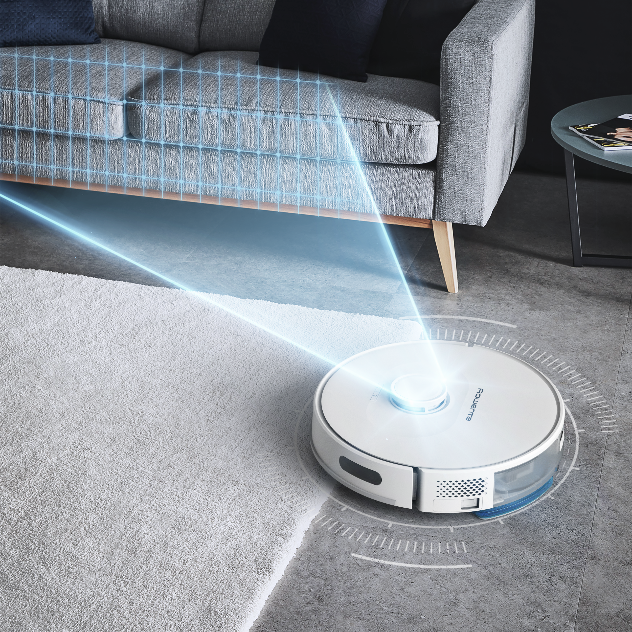 Egy fehér robotporszívó világosszürke szőnyeget tisztít, a háttérben egy kanapé.