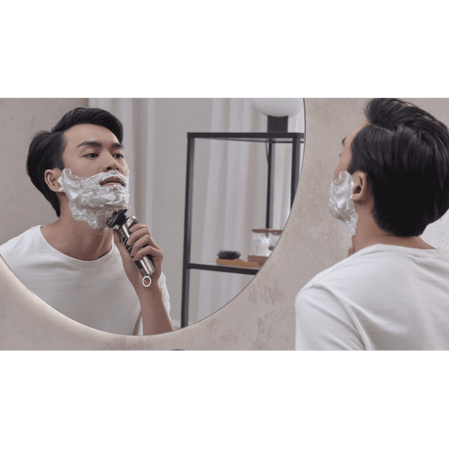 Un homme se rase devant un miroir. Il a de la crème à raser sur le visage et tient un rasoir.