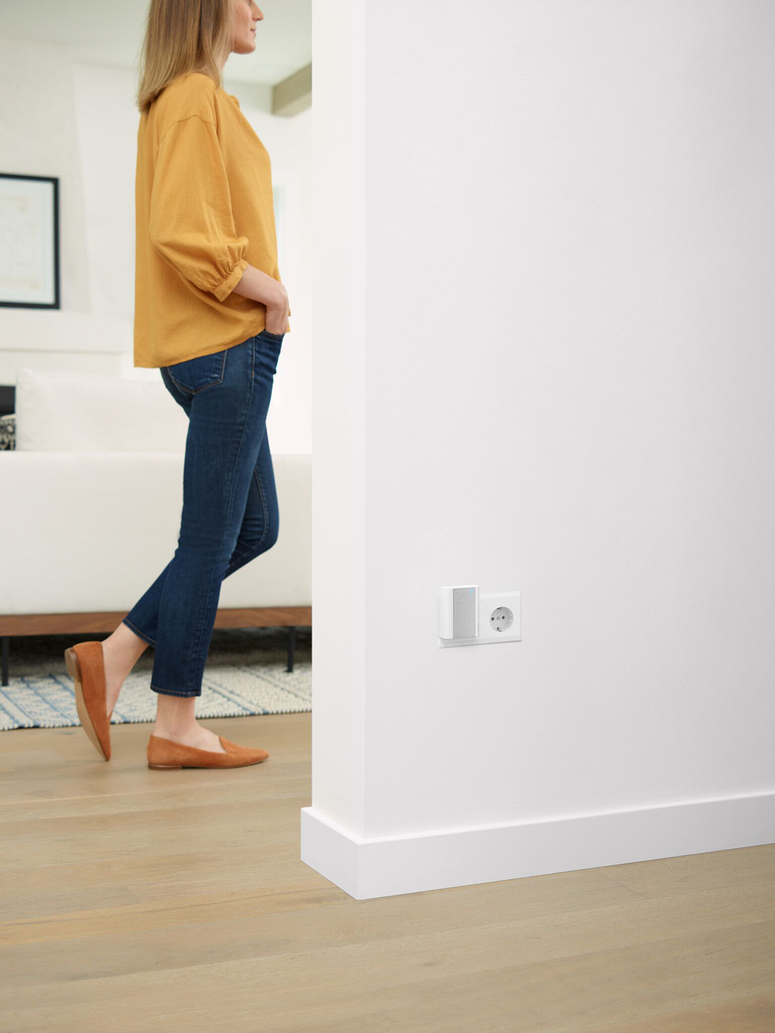 Vrouw in gele top en spijkerbroek loopt langs witte muur met stopcontact en klein wit apparaat.