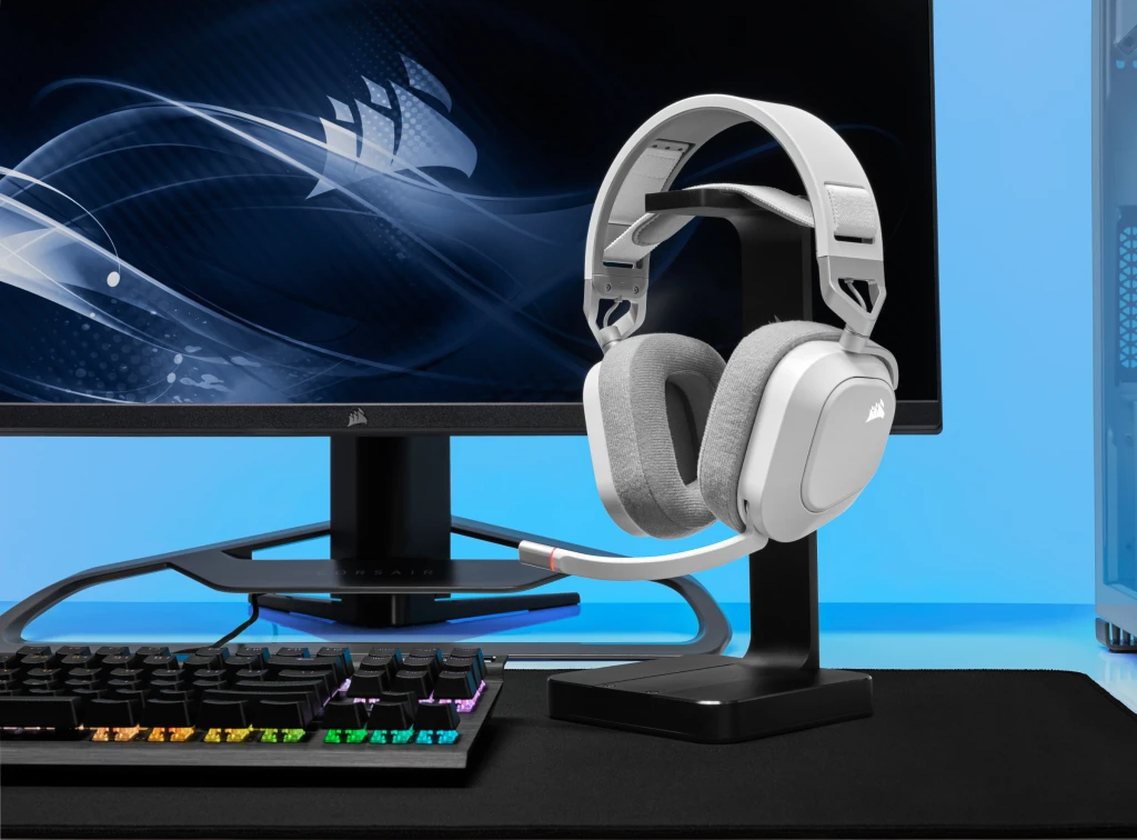 Fehér és szürke gamer headset egy állványon, monitor és billentyűzet előtt.