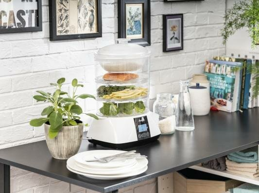 Parowar z jedzeniem w przegródkach, na stole z innymi artykułami kuchennymi.