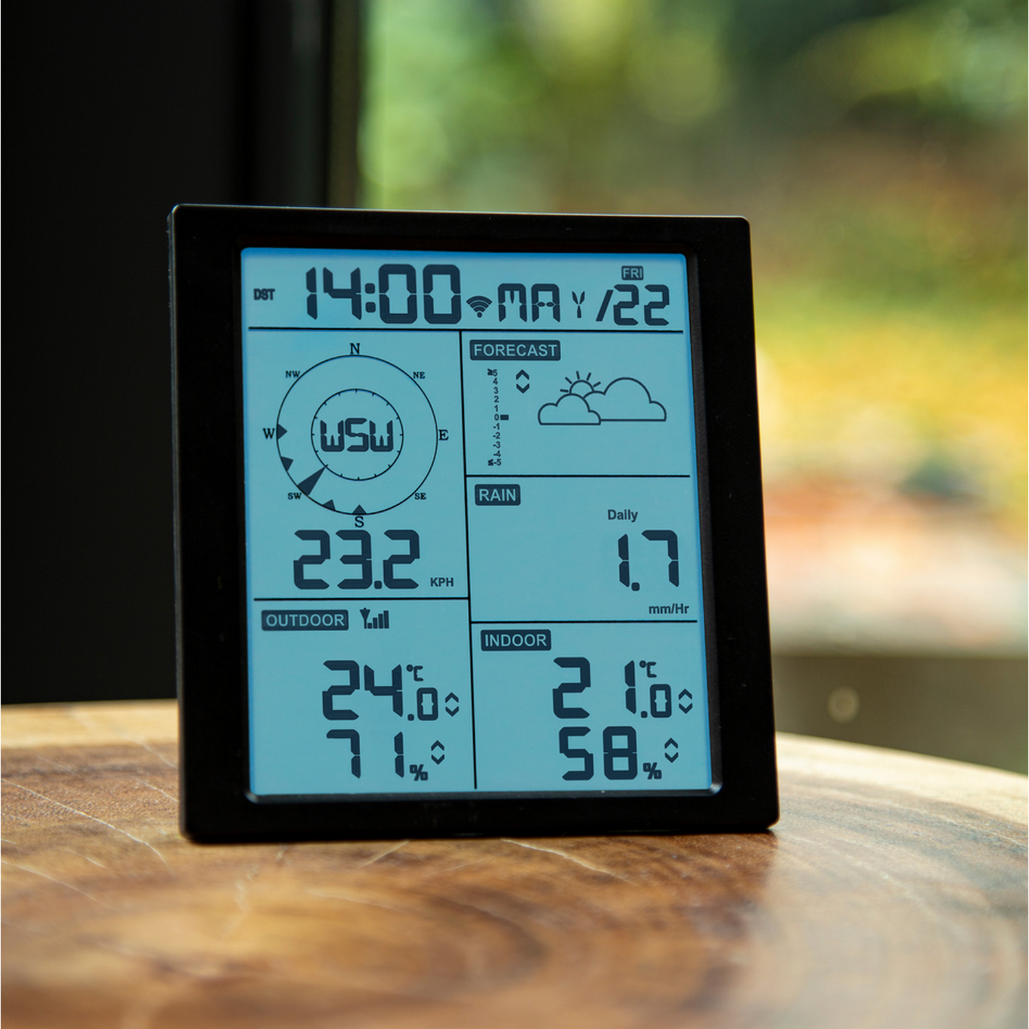 Black digital weather station displaying time, forecast, and temperature readings against a window.