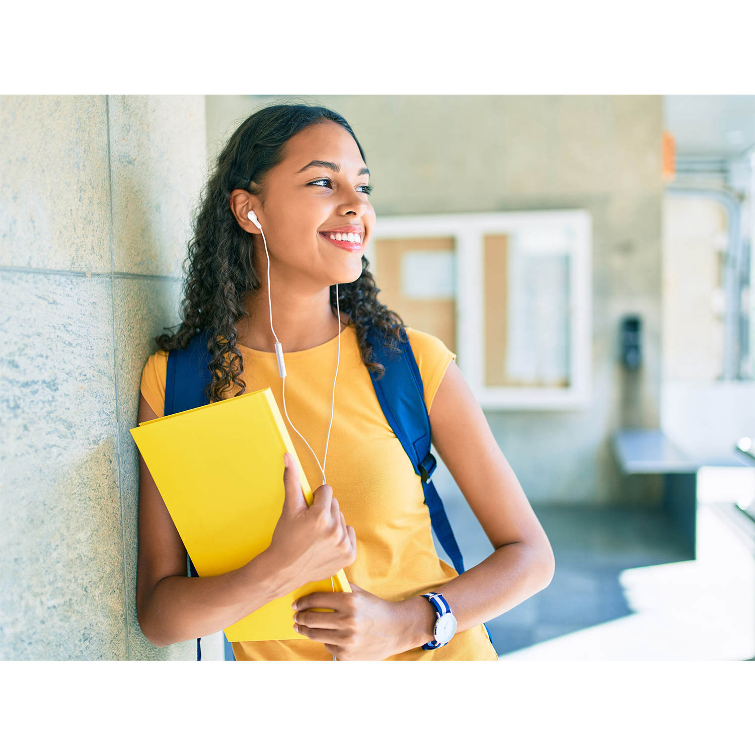 Giovane donna con cuffie e un libro giallo, appoggiata a un muro.