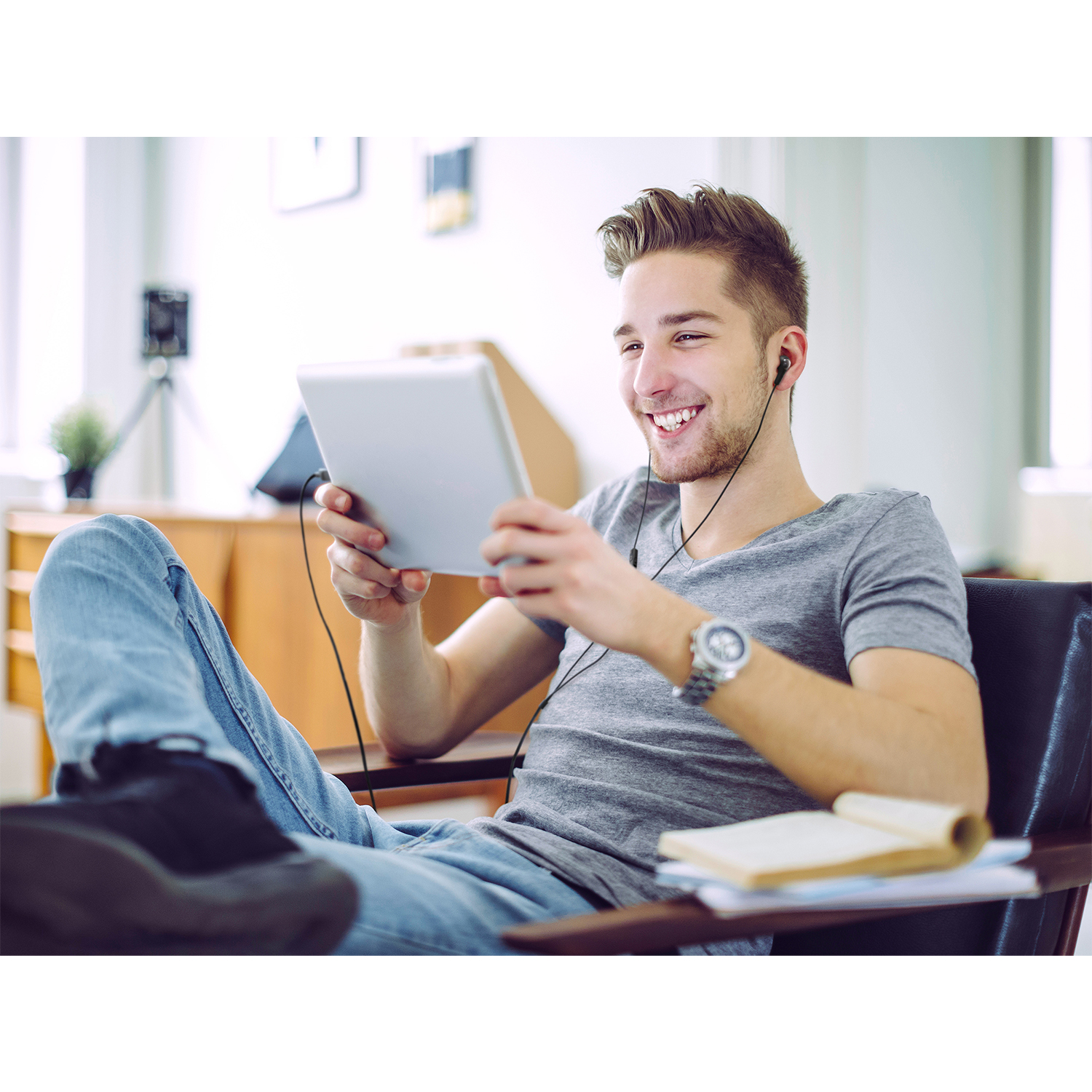 Un giovane sorridente con auricolari, che tiene un tablet, seduto su una sedia.