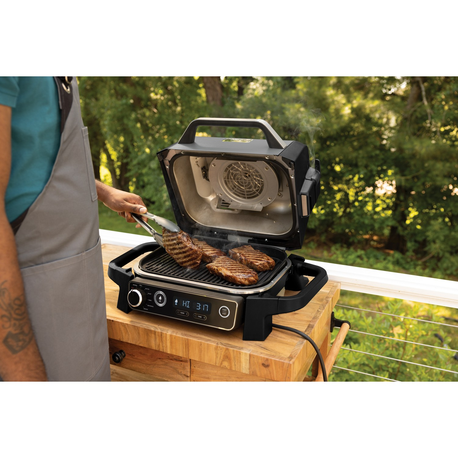 Une personne grillant des steaks sur un barbecue extérieur. Il y a une table en bois et un fond vert.