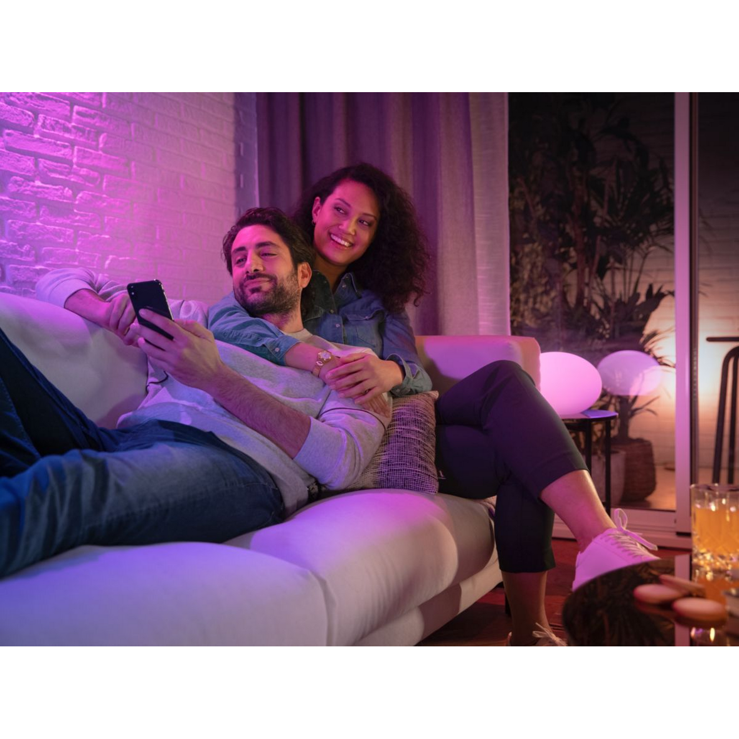 Pareja en sofá blanco, hombre usando teléfono, mujer sonriendo. Luces rosas y blancas al fondo.