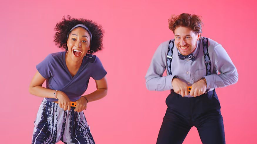 Due persone che tengono dei bastoni arancioni, sorridendo. Sfondo rosa.