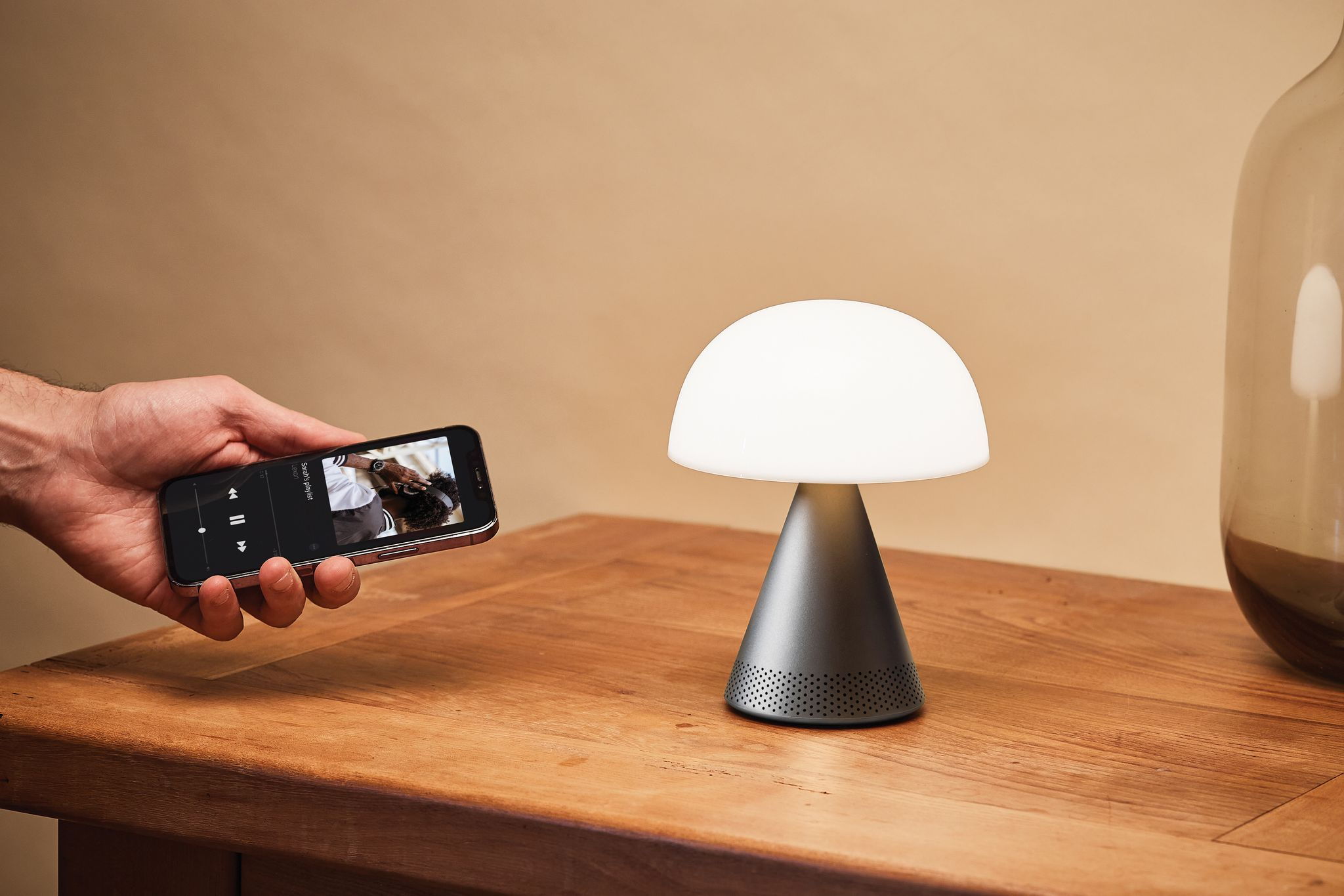 Une personne tenant un smartphone à côté d'une lampe en forme de champignon. Table en bois.